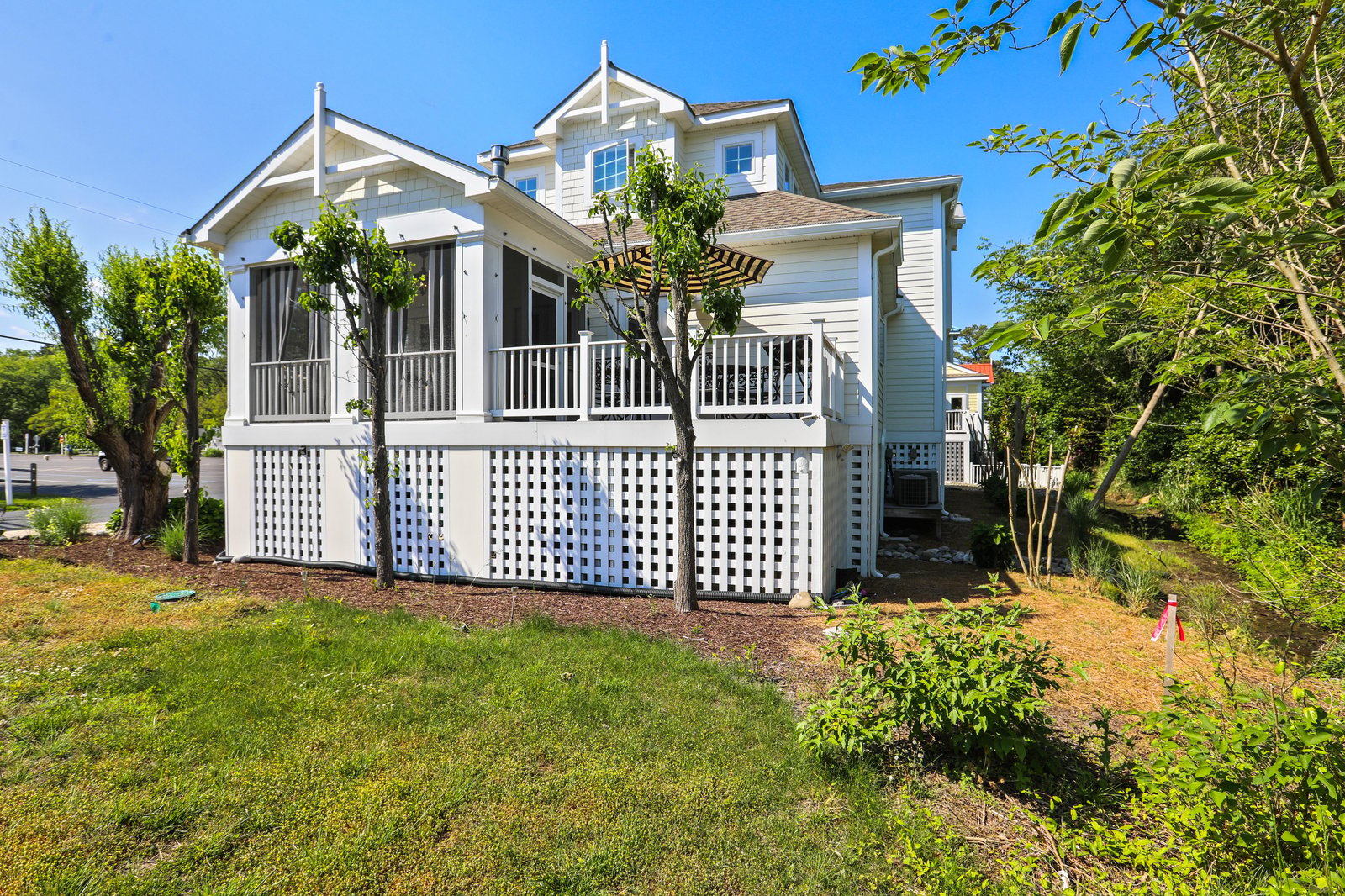 A charming coastal-style property with classic white lattice detailing and inviting wraparound porches, nestled in a peaceful, tree-lined neighborhood under sunny skies.