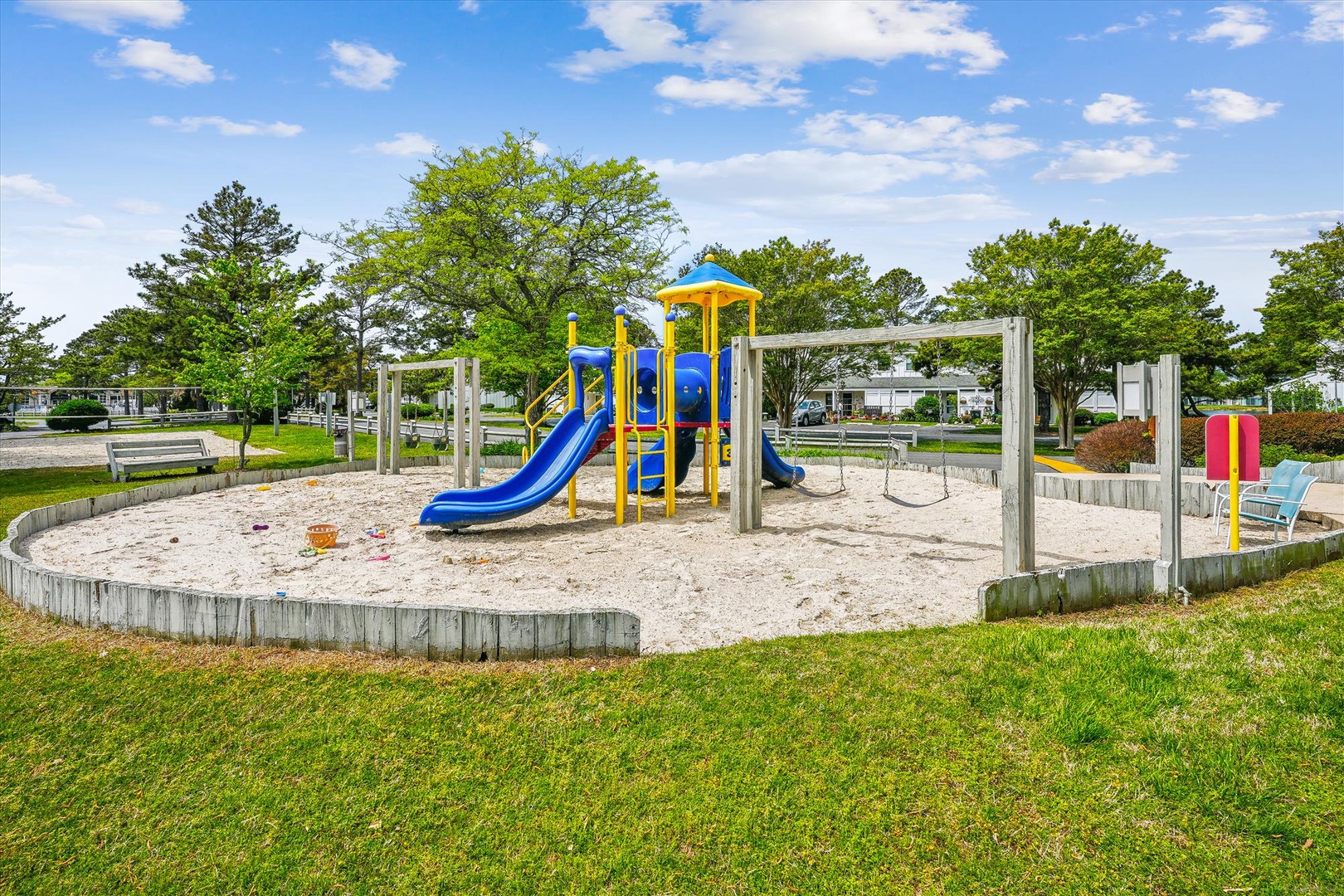 Colorful playground with slides and swings set in a sandy play area, surrounded by lush green spaces and mature trees.