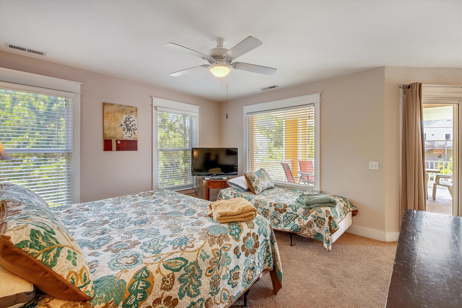 Your spacious bedroom offers two plush beds dressed in tropical linens, a wall-mounted TV, ceiling fan for comfort, and bright windows revealing leafy views and balcony access.