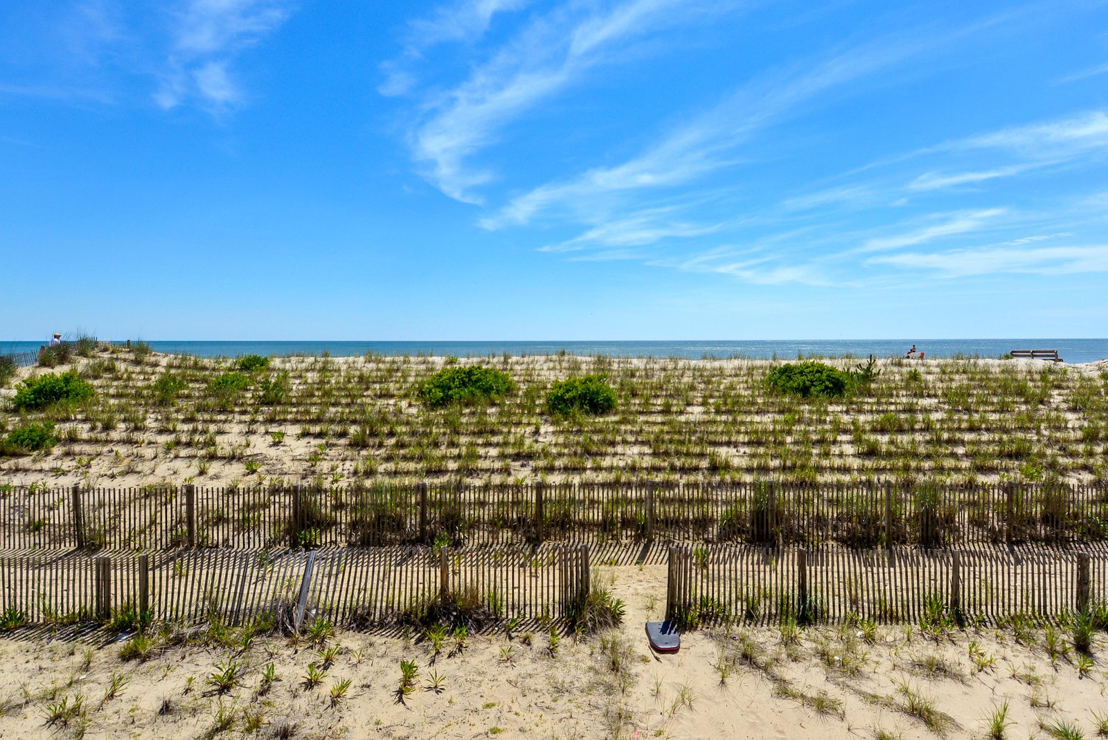 A pristine sandy beach unfolds beneath a brilliant blue sky, bordered by natural dune grasses and fencing that protect the coastal landscape.