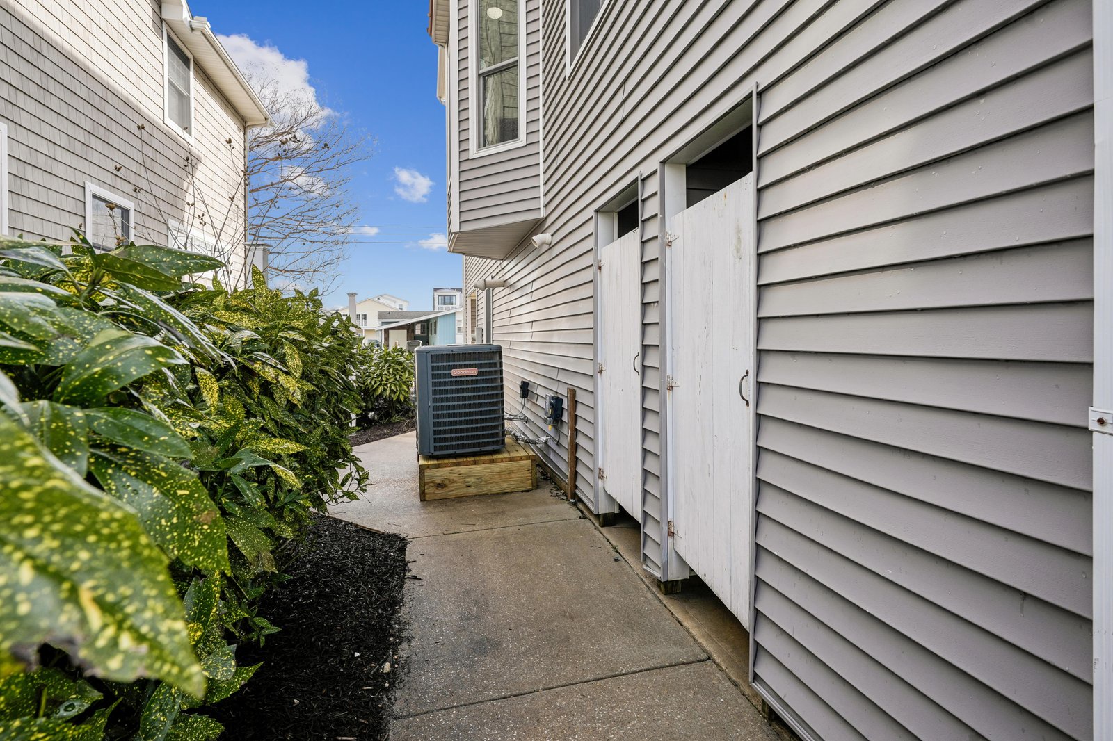 A narrow exterior walkway between buildings with green landscaping and an air conditioning unit visible along the property's side access.