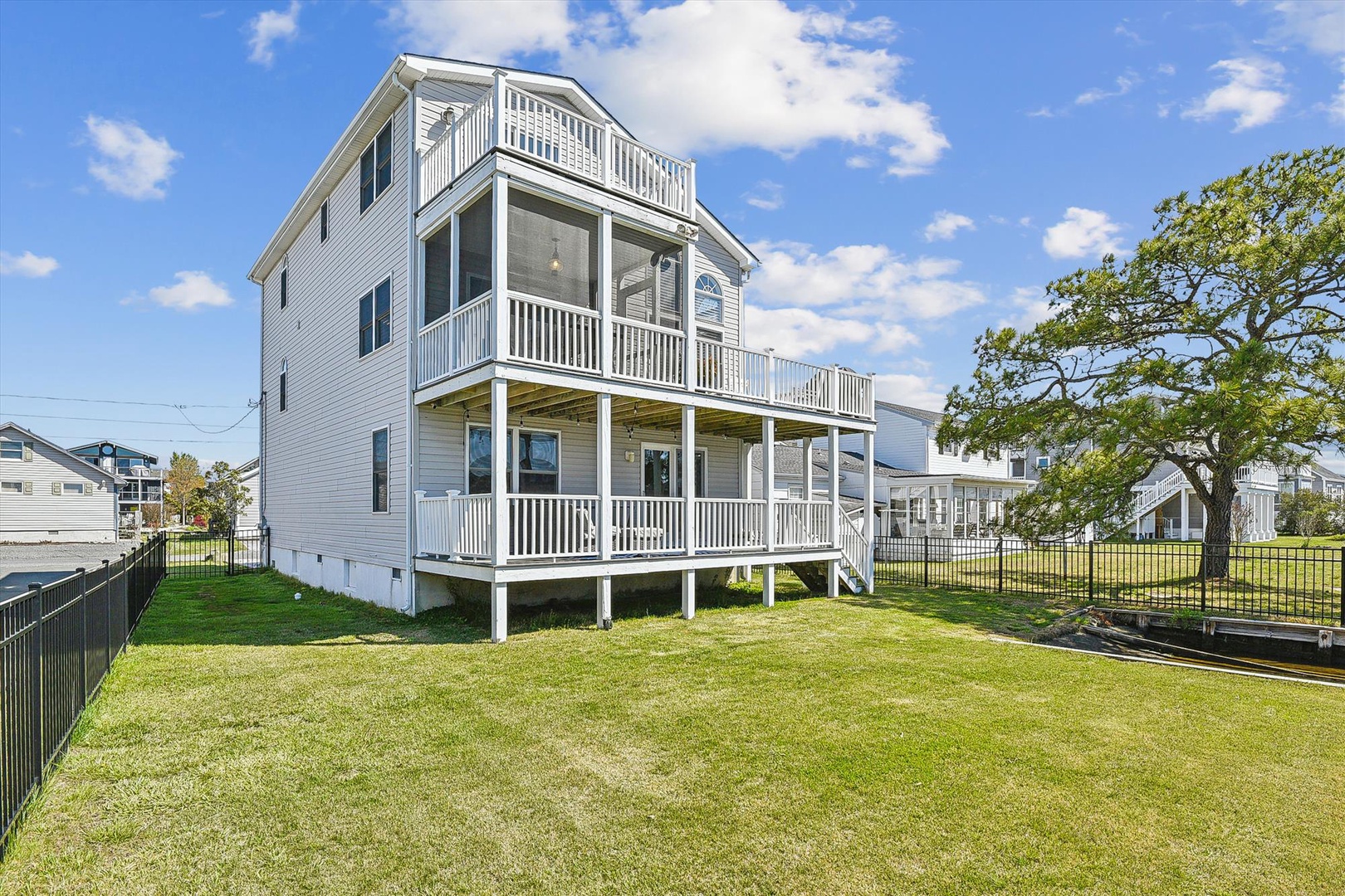 A spacious three-story coastal home with expansive porches on each level, surrounded by a manicured lawn and mature shade tree. The white exterior and multiple balconies create a classic beach-house aesthetic in this quiet residential neighborhood.