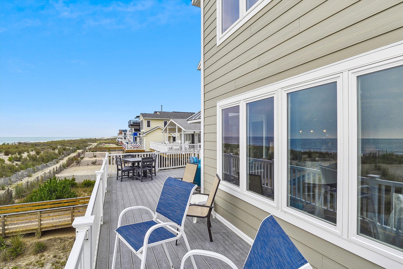 Beachfront property featuring multiple levels of outdoor decking with direct ocean access, surrounded by coastal dunes and neighboring vacation homes under clear blue skies.