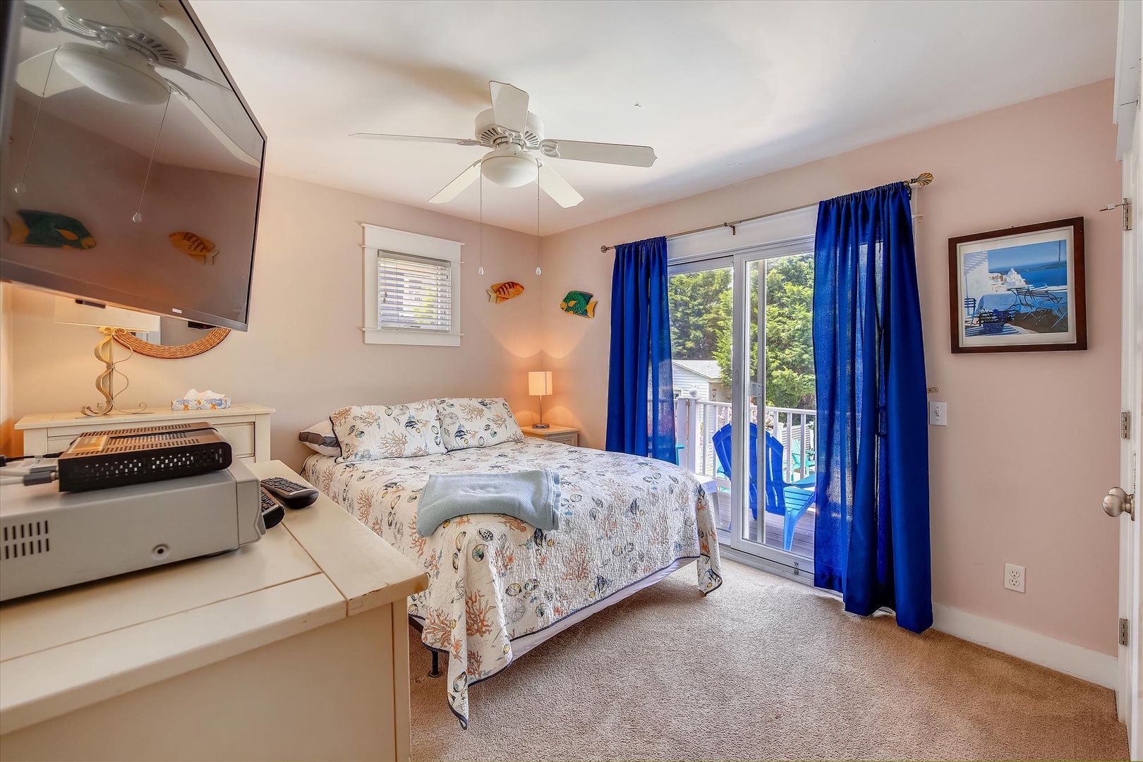 Step into your coastal retreat, where ocean-themed décor and breezy blue curtains frame your private balcony access. The ceiling fan keeps you cool as you unwind in this cheerful, light-filled bedroom.