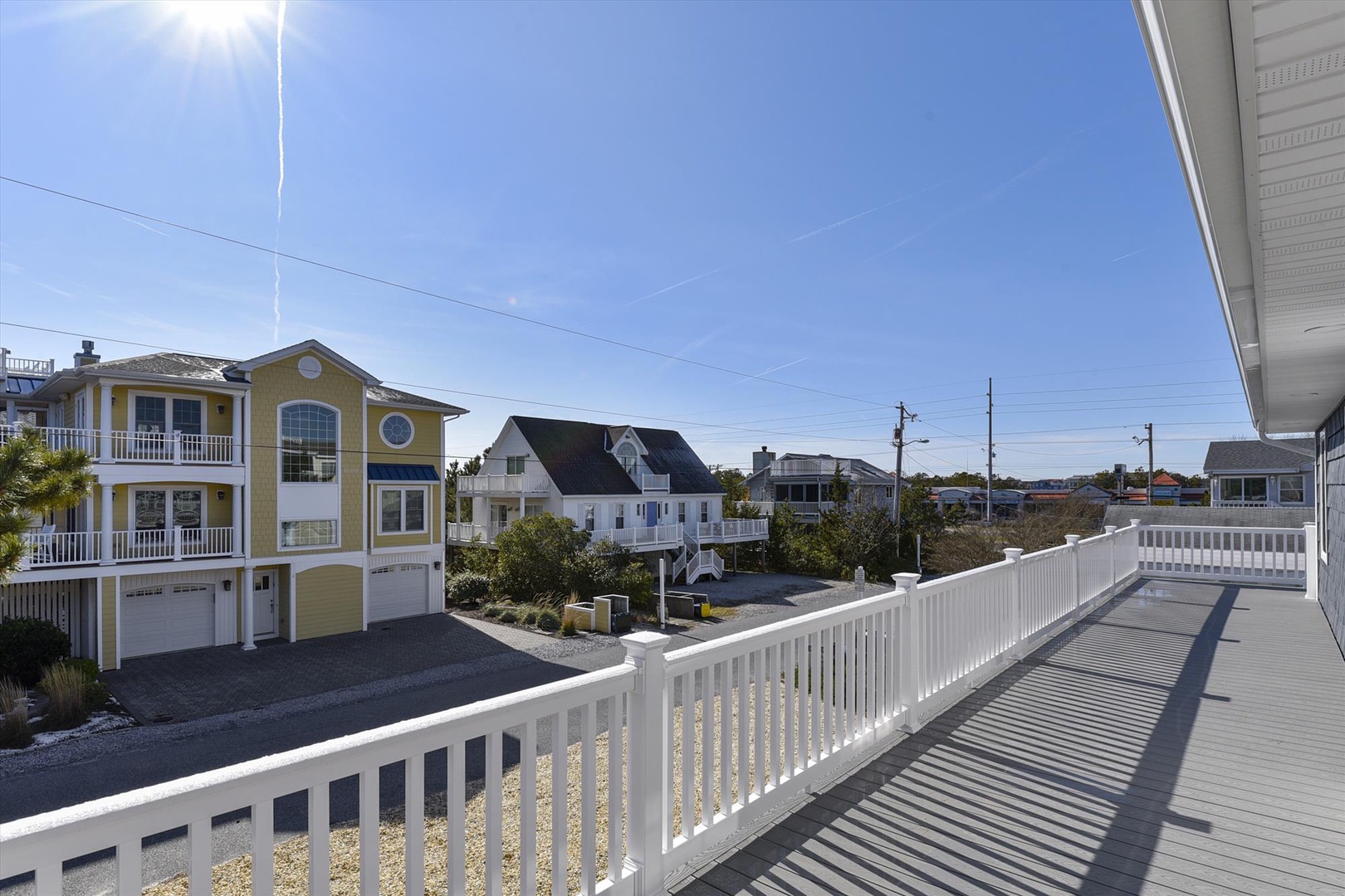 Spacious deck featuring a white railing overlooks a residential coastal neighborhood with beach homes and clear blue skies, offering a peaceful setting near the shore.