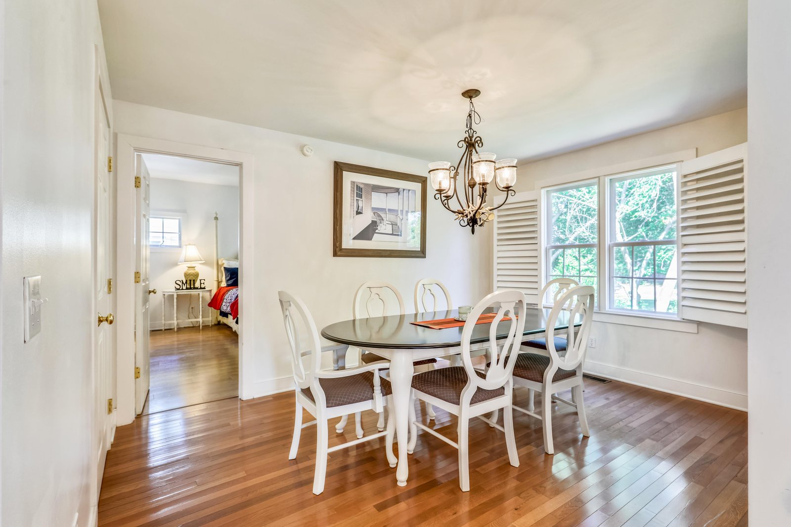 Gather around this inviting dining table beneath a warm chandelier, with leafy views through plantation shutters and polished hardwood underfoot creating a perfect spot for your meals together.