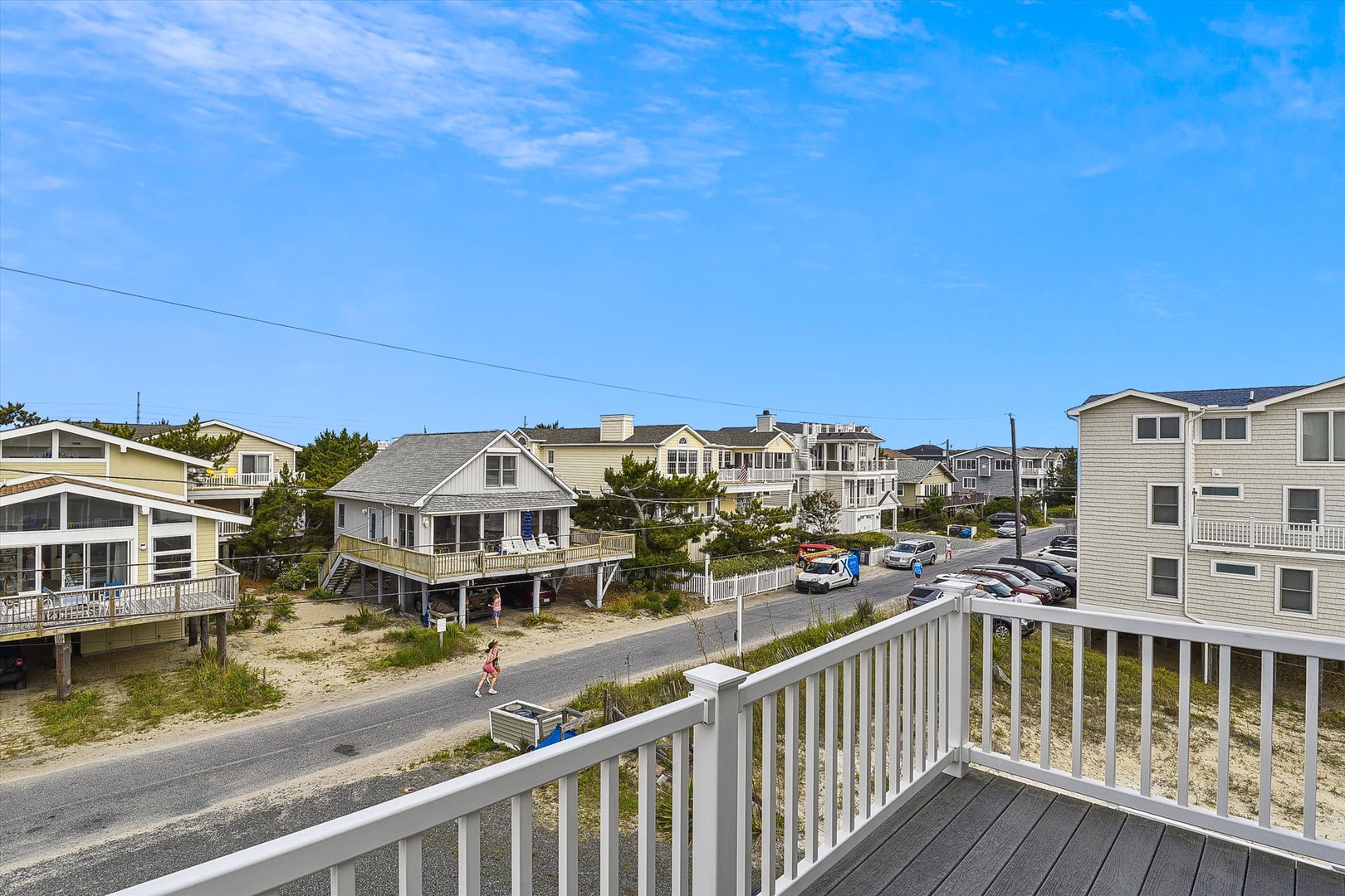 A spacious balcony overlooking a quiet beach neighborhood, framed by coastal homes and sandy dunes under clear skies.