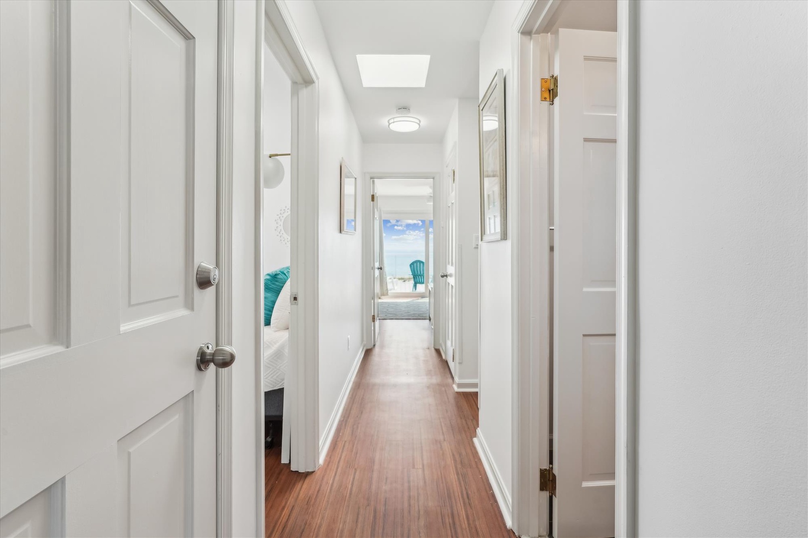 A sunlit hallway draws your eye through crisp white doors and warm wood floors to a stunning sea view at the end, promising coastal tranquility throughout your stay.