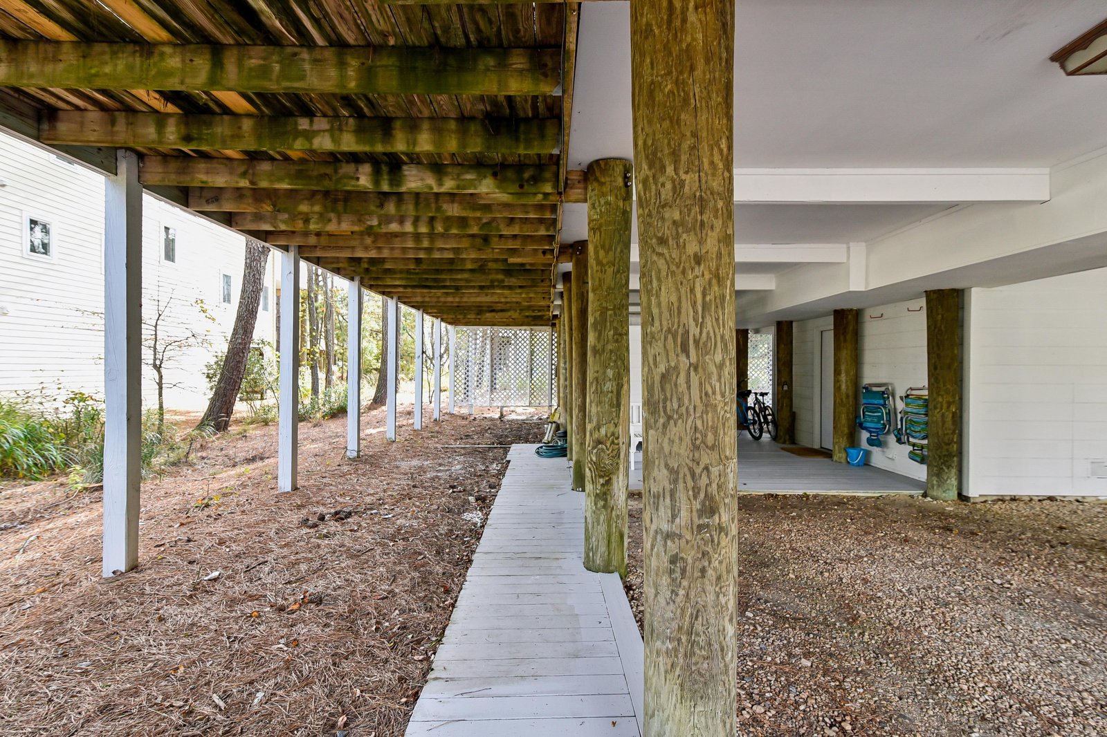 A covered walkway wraps beneath the property, showcasing the raised foundation, sturdy wooden beams, and gravel parking area—perfectly suited for beach living and coastal weather.