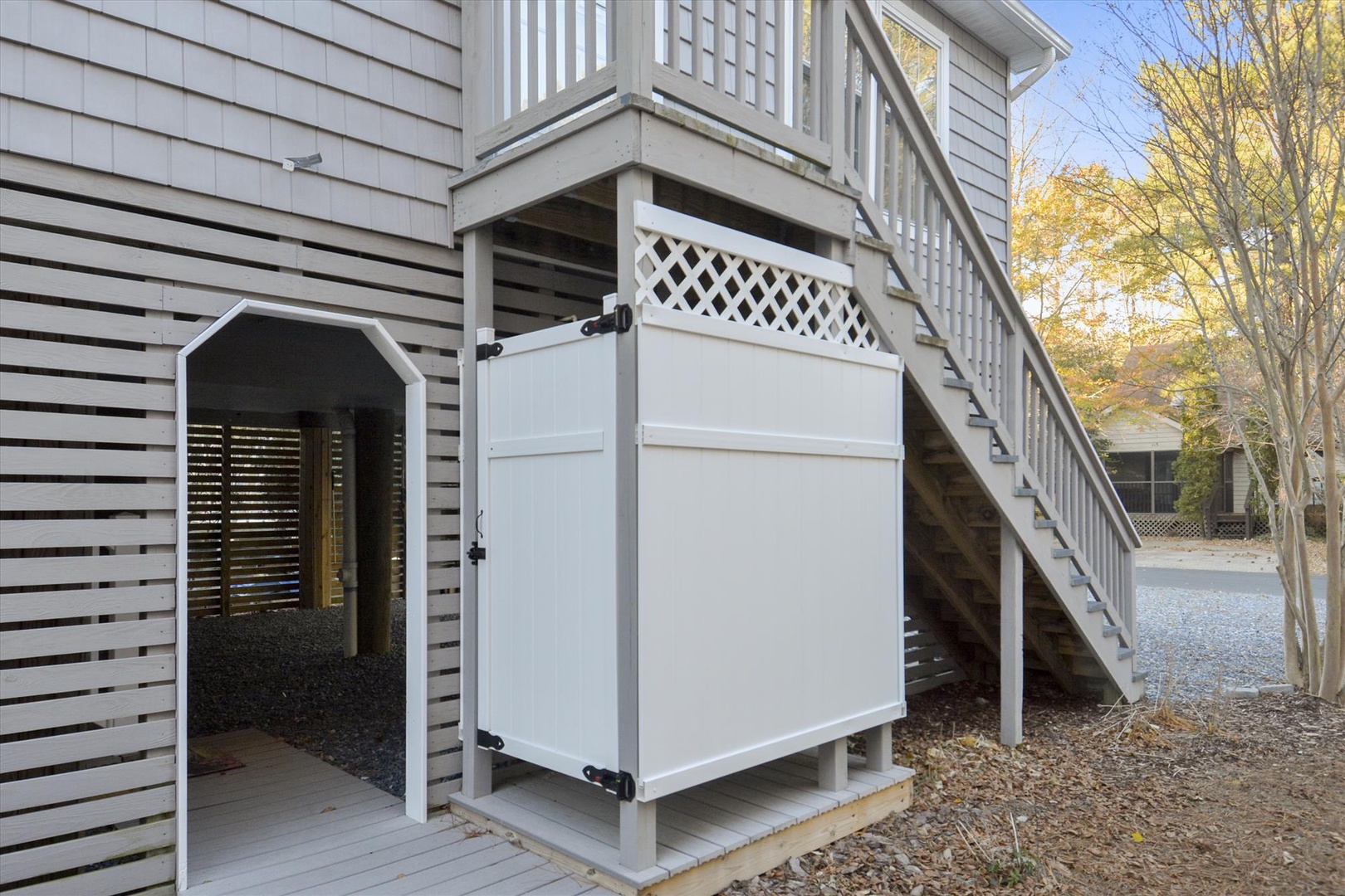 A charming exterior entry features a practical enclosed storage area beneath elevated stairs, with mature trees displaying autumn foliage in the background.