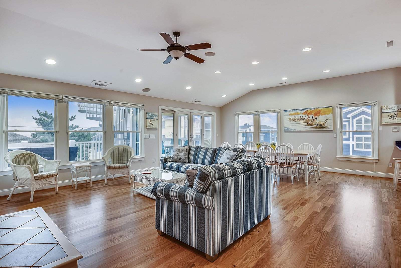 Spacious, sun-soaked living area with gleaming hardwood floors and coastal charm. Relax on striped sofas, share meals at the dining table, and enjoy ocean breezes through the windows.