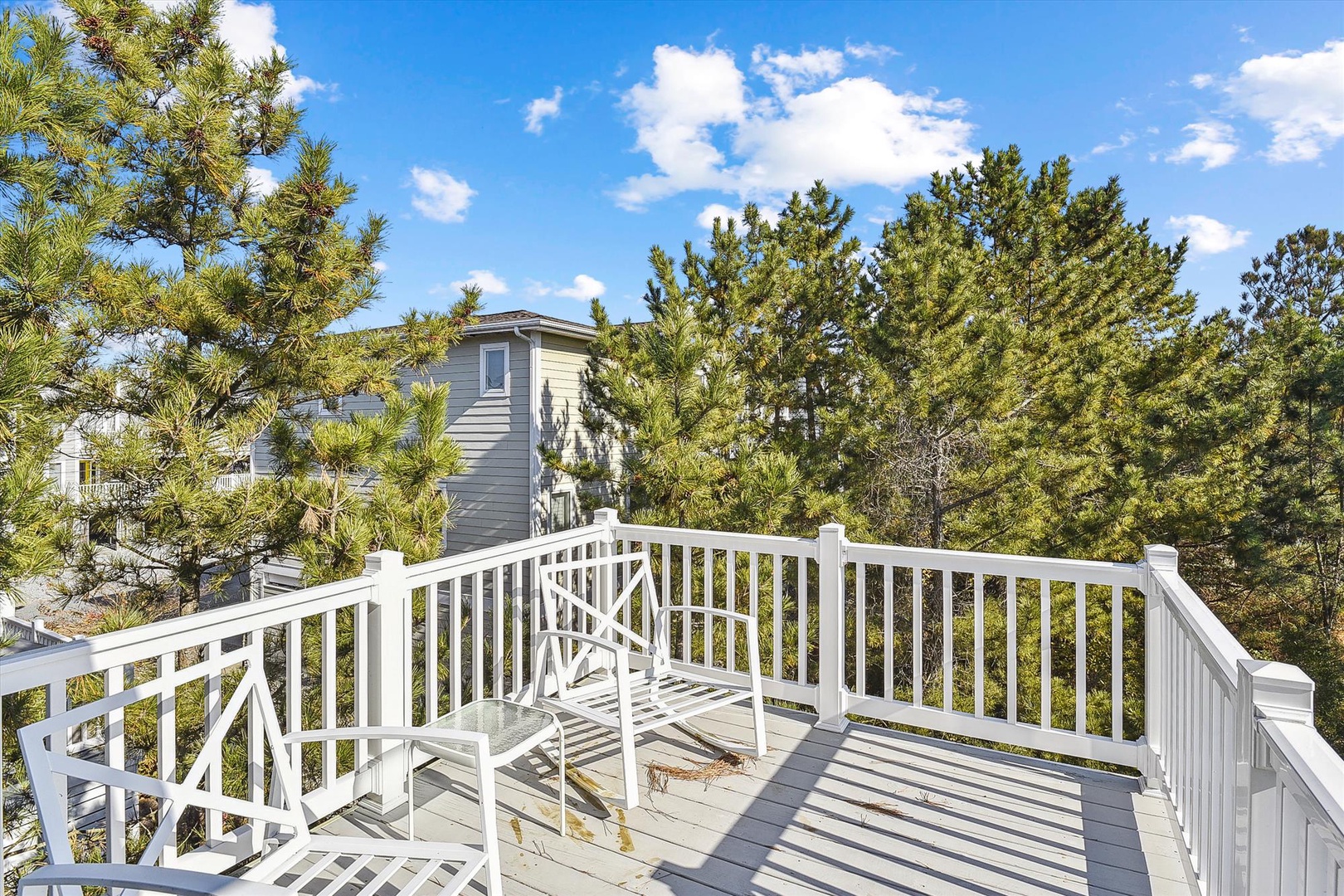 A spacious outdoor deck framed by tall pines, offering an elevated vantage point overlooking the neighborhood under a bright blue sky.