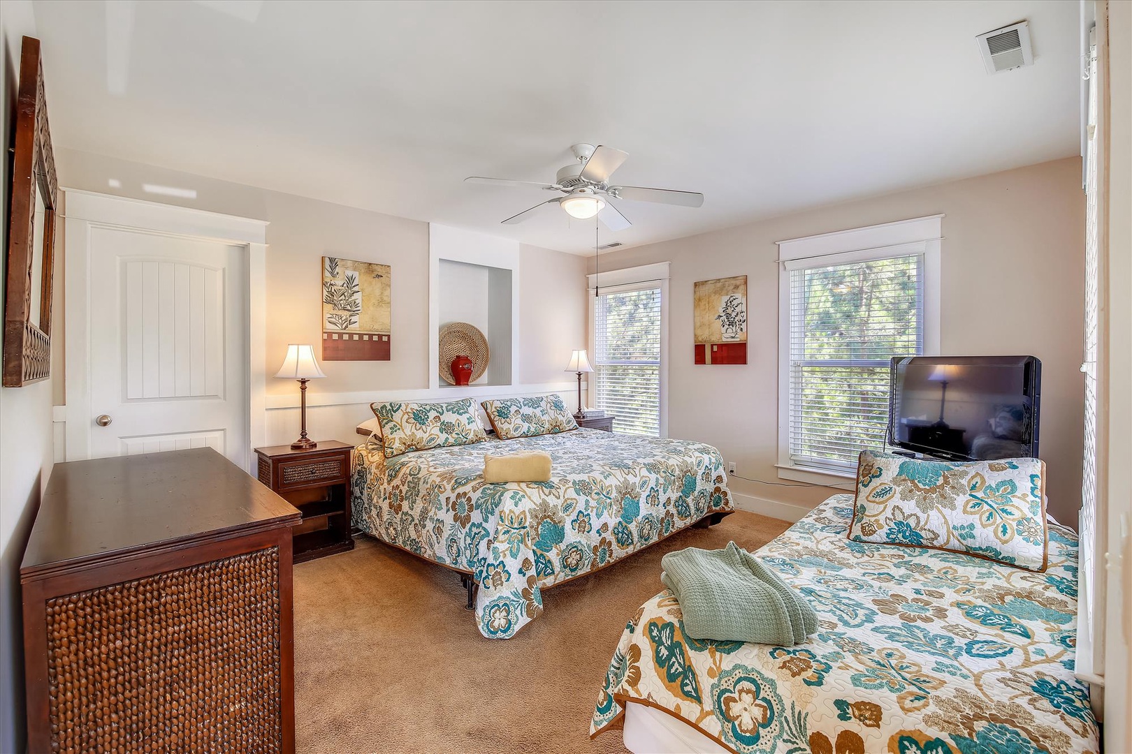 Your spacious bedroom features comfortable beds with vibrant linens, warm bedside lighting, a ceiling fan, flat-screen TV, and bright windows overlooking natural greenery—perfect for restful mornings and peaceful nights.