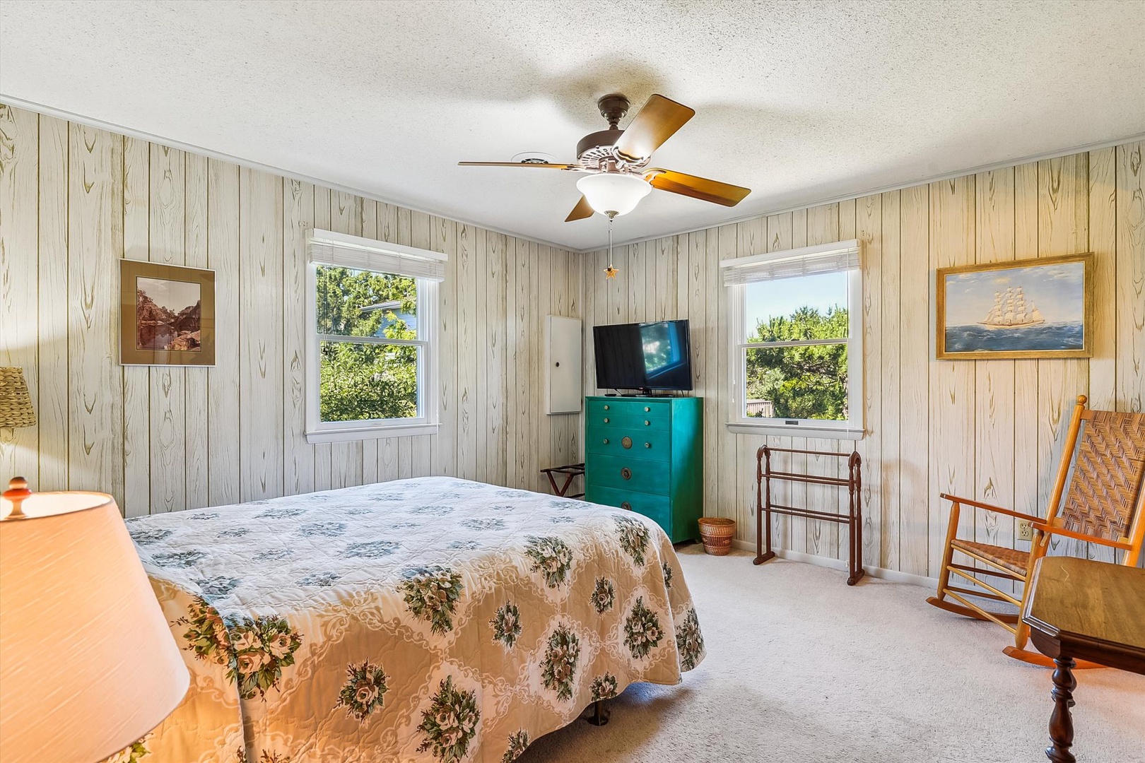 Relax in your bright, airy bedroom with coastal charm. Gentle breezes from the ceiling fan, twin windows framing lush greenery, and a cozy rocking chair await your peaceful retreat.