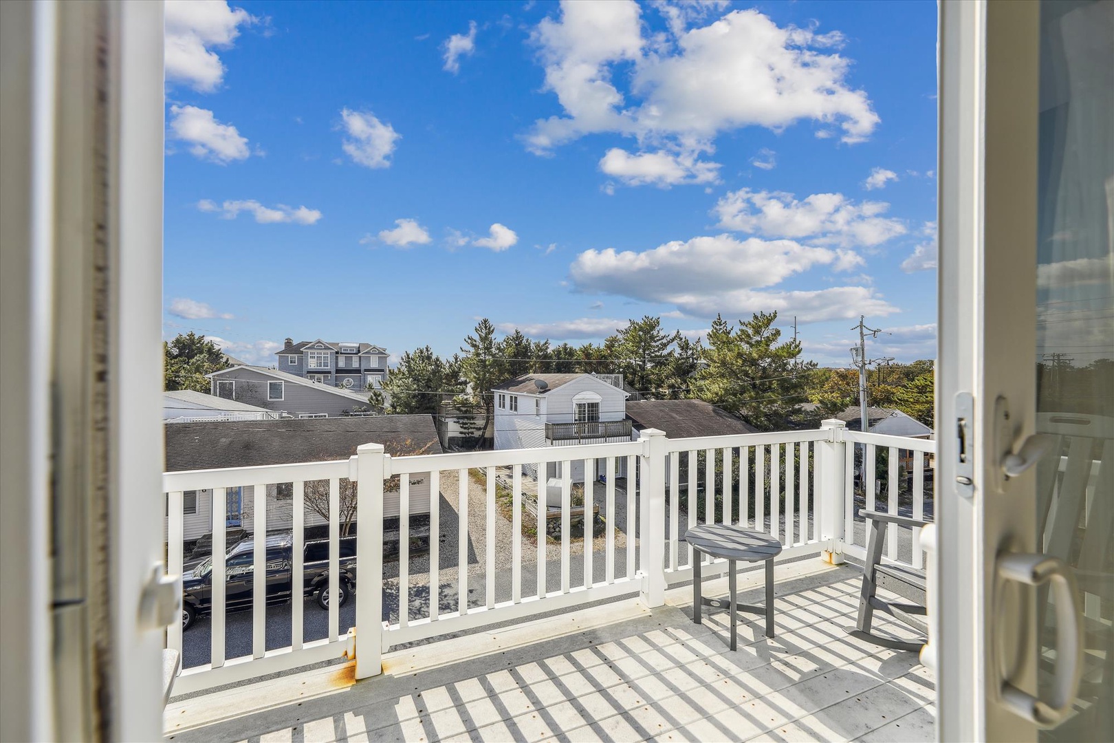 A private balcony with seating overlooks a quiet residential neighborhood lined with trees and coastal homes under open skies.