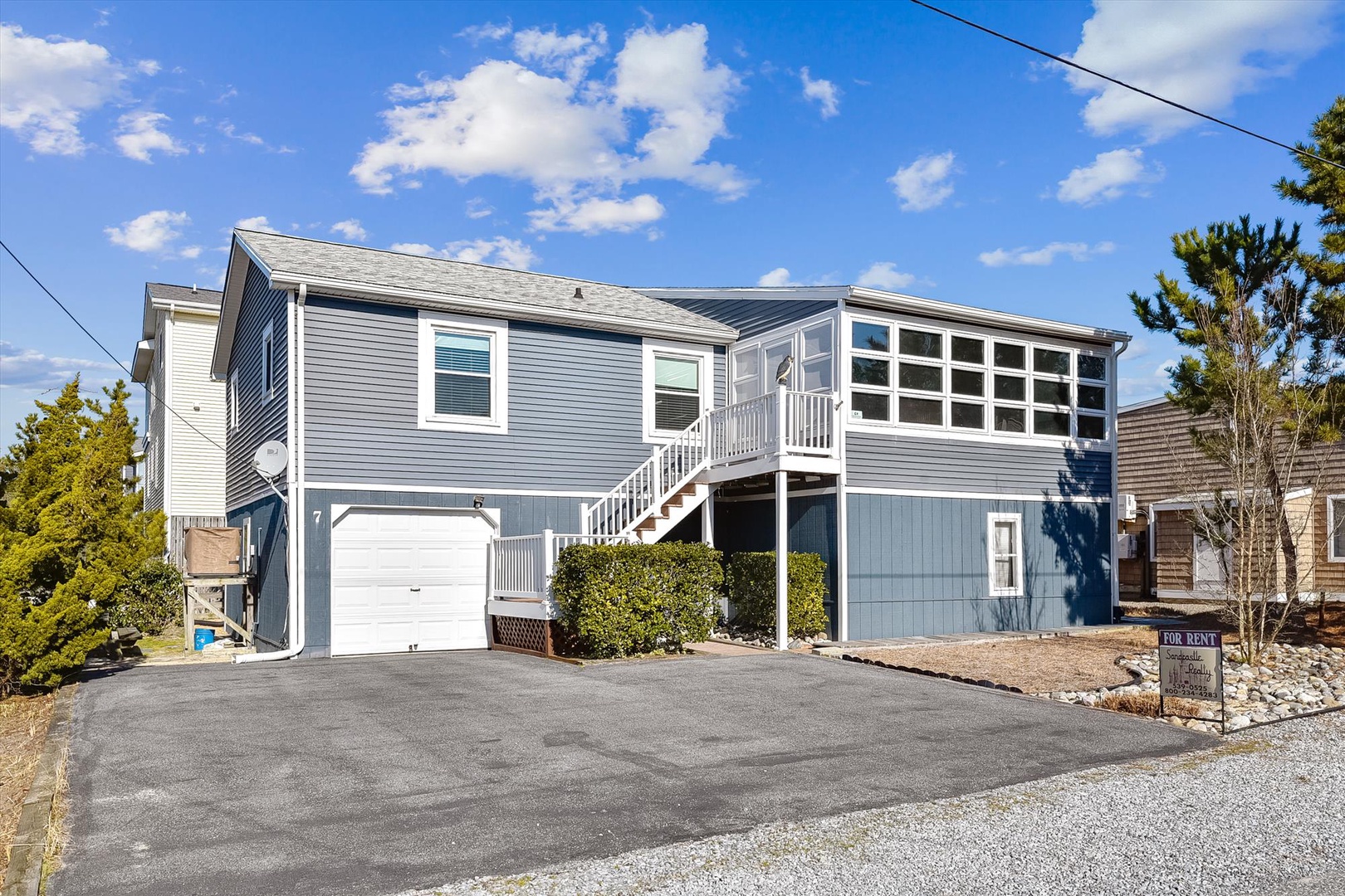 A charming blue coastal home with enclosed upper deck offers private parking and easy access, situated in a quiet residential setting surrounded by greenery.
