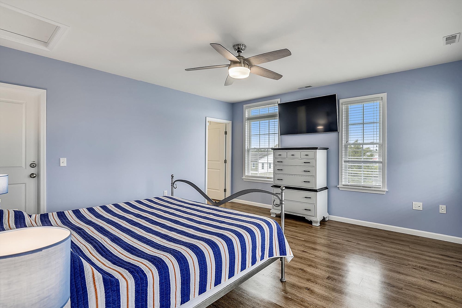 Your spacious bedroom features soft blue walls, natural light from dual windows, and a ceiling fan for your comfort. The bright, airy space includes a mounted TV and ample storage, creating a peaceful coastal-inspired retreat for restful nights.