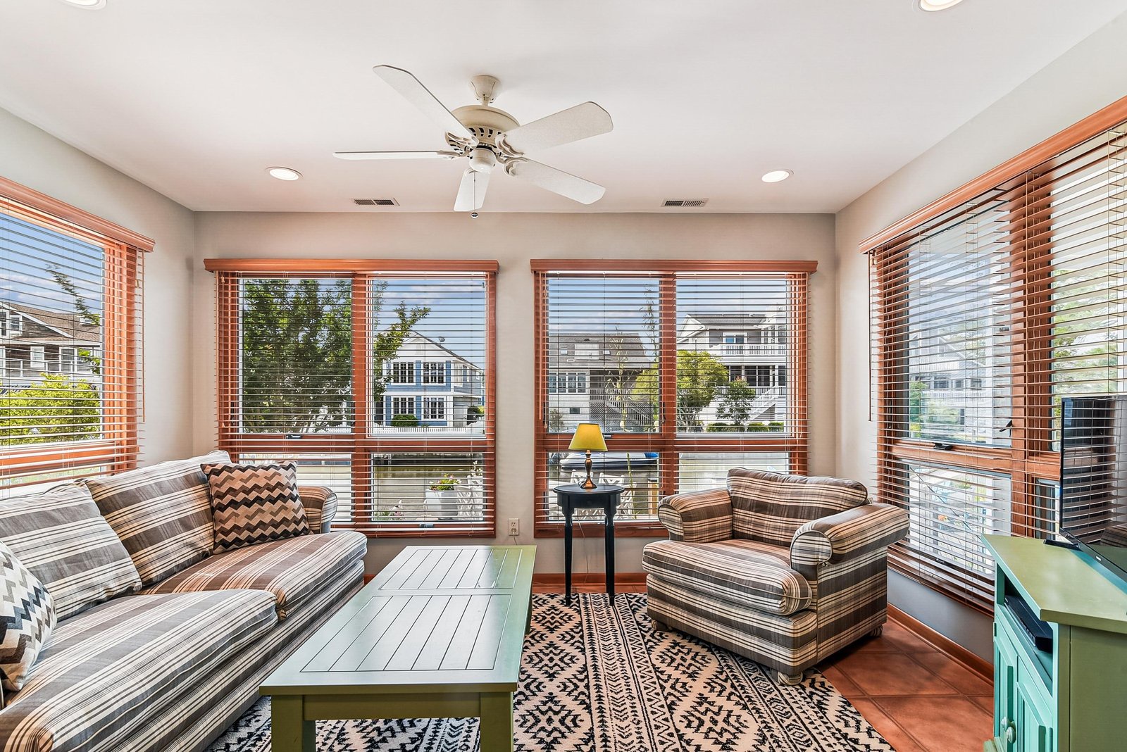 Relax in your bright sunroom with stylish seating and a gentle ceiling fan, while warm wood blinds frame the sunny day outside.