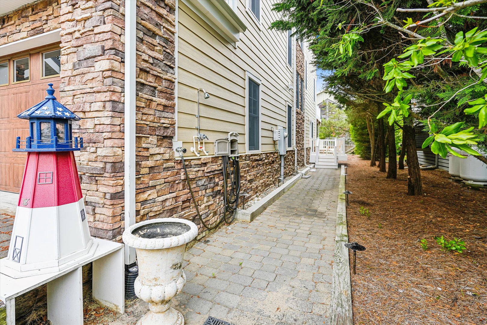 Charming coastal entrance marked by a decorative red-and-white lighthouse, with stone and siding architecture set among mature trees and a tranquil, shaded pathway leading to the property.