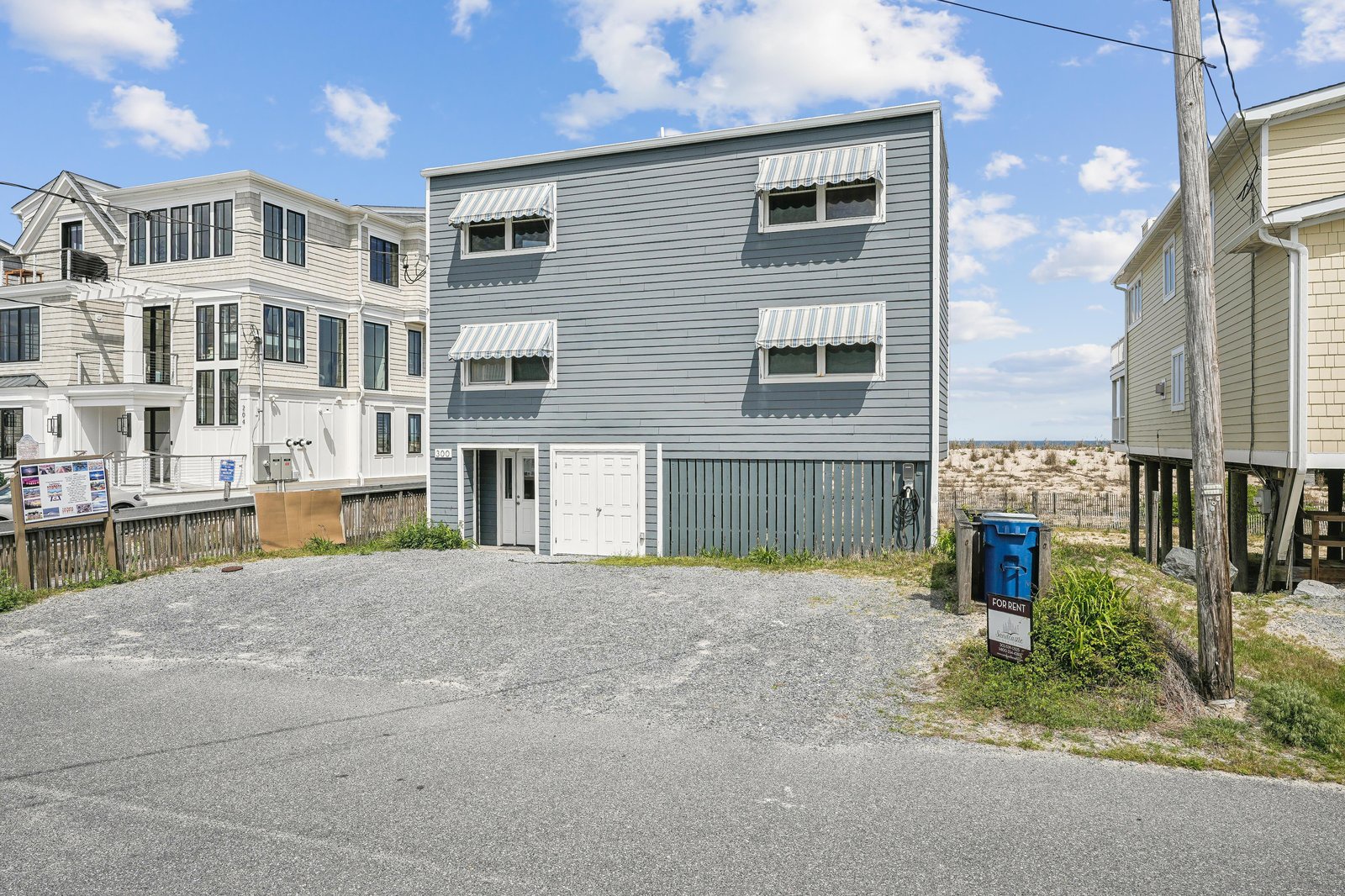 A coastal three-story beach house with striped awnings nestled among neighboring homes, offering a quiet neighborhood setting just steps from the shore.