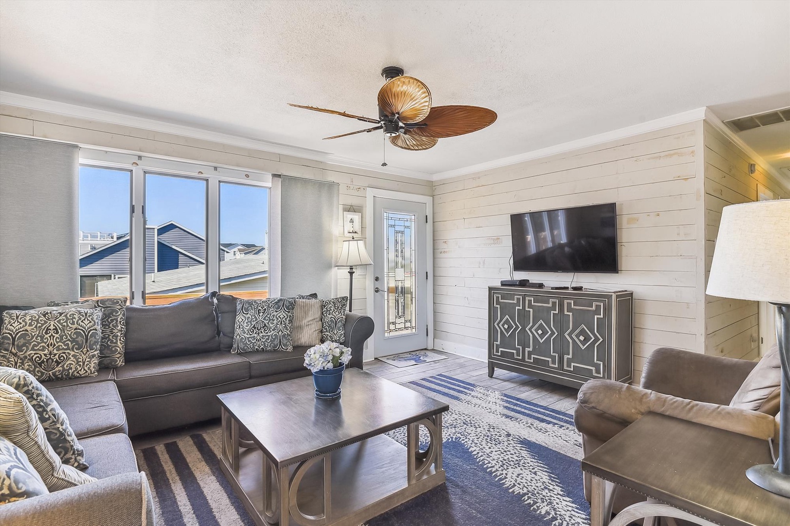 Relax in your bright, coastal-inspired living room with plush seating, tropical ceiling fan, and flat-screen entertainment. Large windows welcome natural light while shiplap walls create timeless charm.