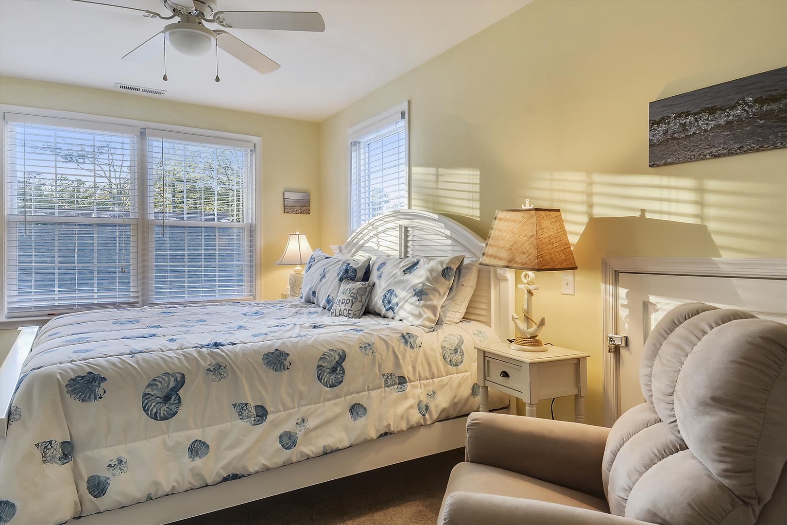 Relax in your coastal-themed bedroom with nautical bedding, soft natural light from large windows, ceiling fan, and a cozy reading chair—your peaceful seaside retreat awaits.