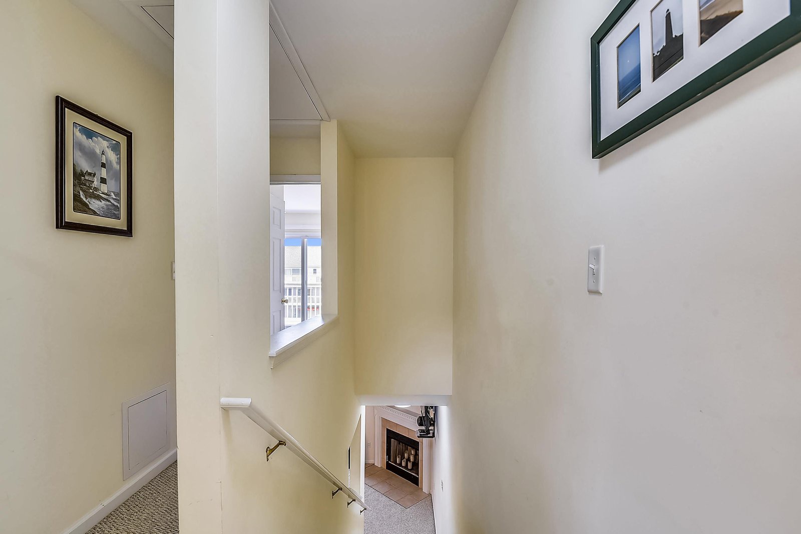 A bright, sun-lit stairway connects your living spaces with a charming window overlooking the surroundings, enhanced by coastal artwork and a cozy fireplace below.
