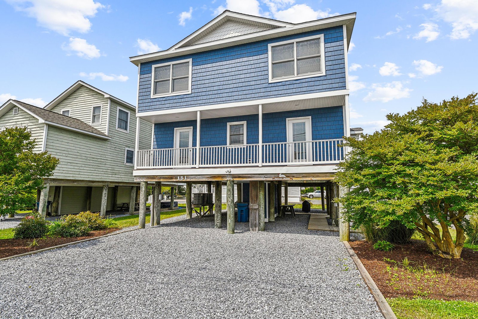 A charming coastal-style home with blue siding sits on stilts, featuring a welcoming second-floor porch and gravel parking beneath. Nestled in a quiet neighborhood with lush greenery surrounding.