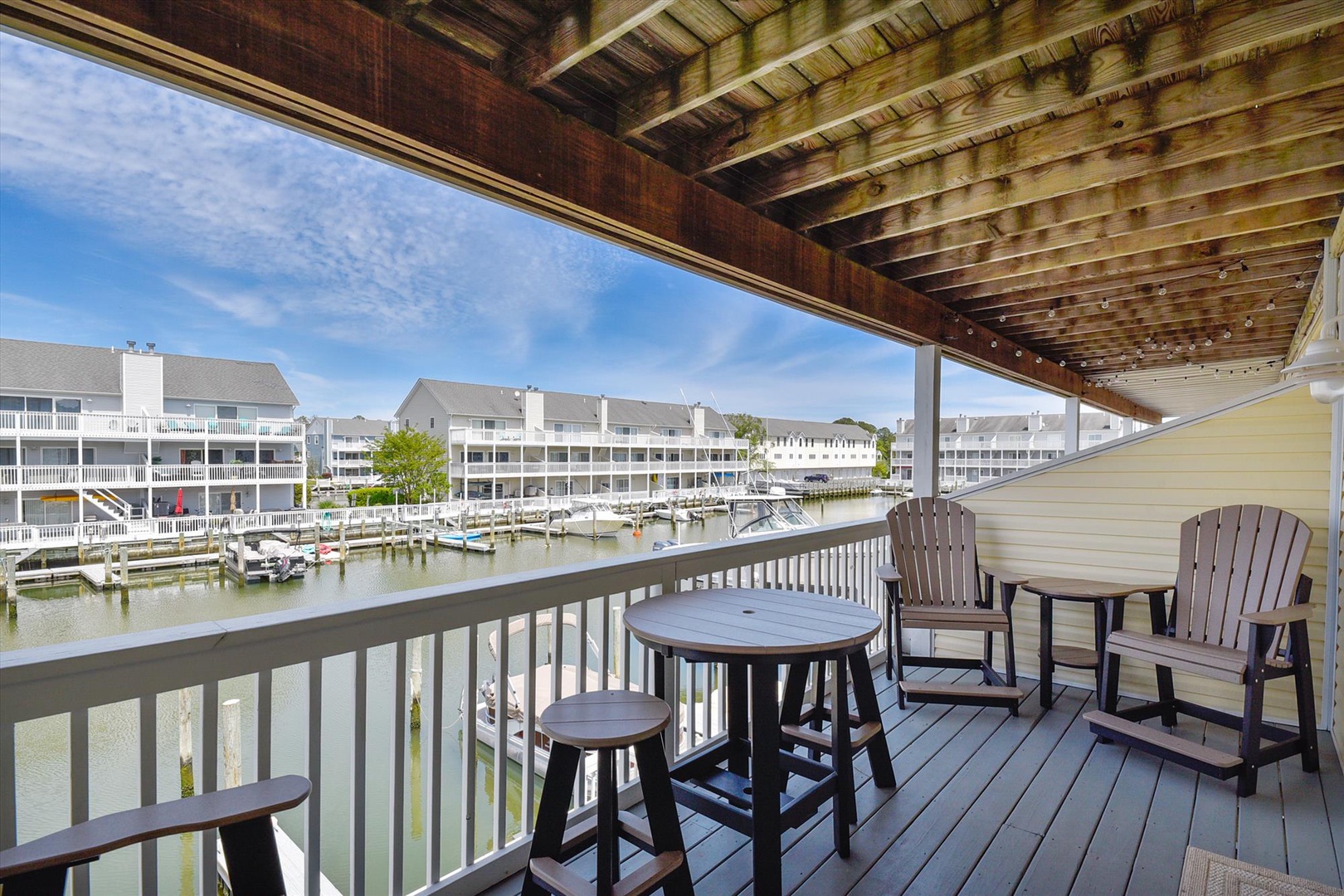 Your private waterfront balcony overlooks a peaceful marina where boats bob gently at their slips. Relax in comfortable Adirondack chairs under the warm wooden pergola, enjoying serene water views.