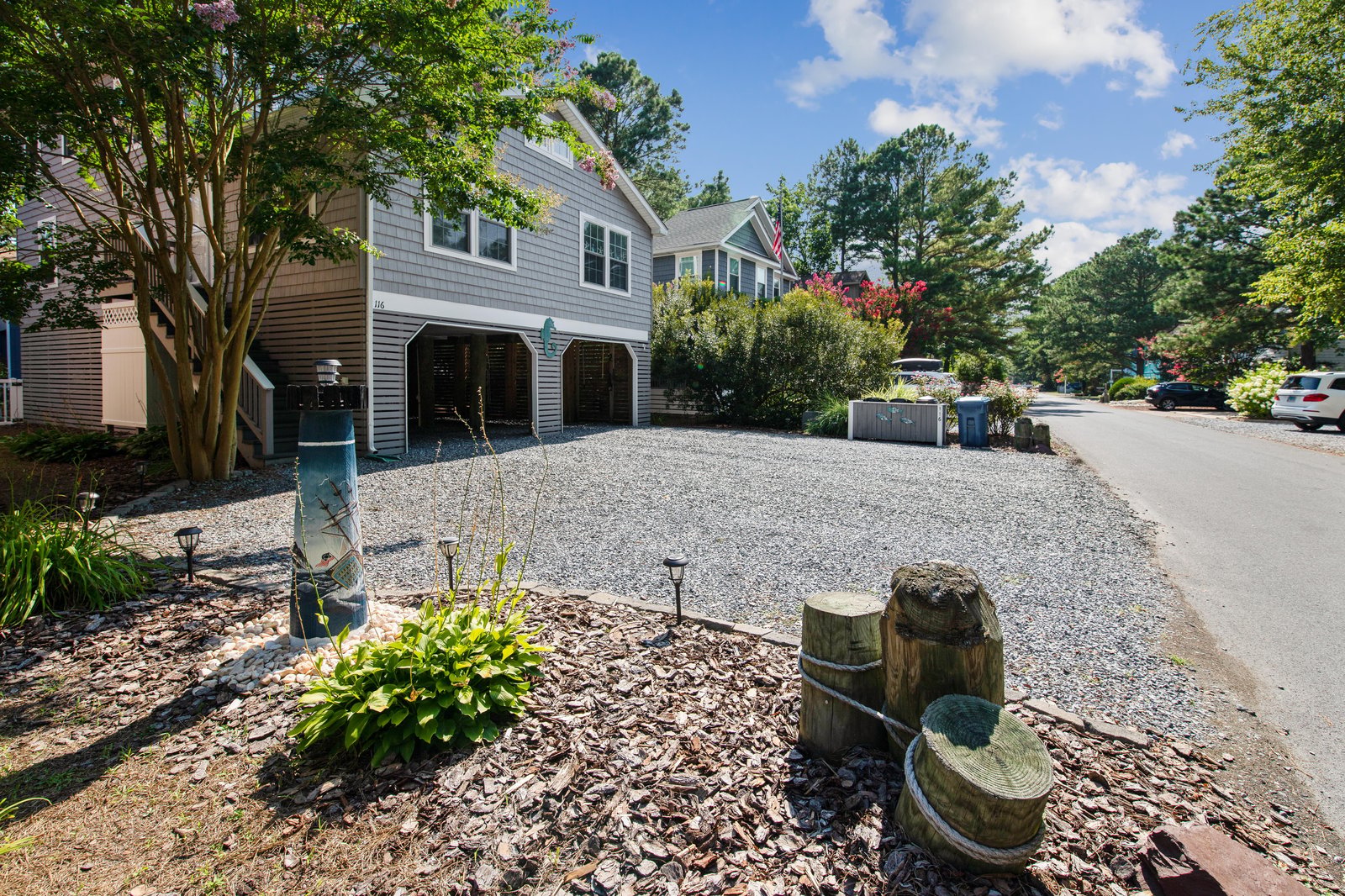 A charming coastal-style property nestled in a lush, tree-lined neighborhood with ample parking and manicured gardens welcoming guests upon arrival.