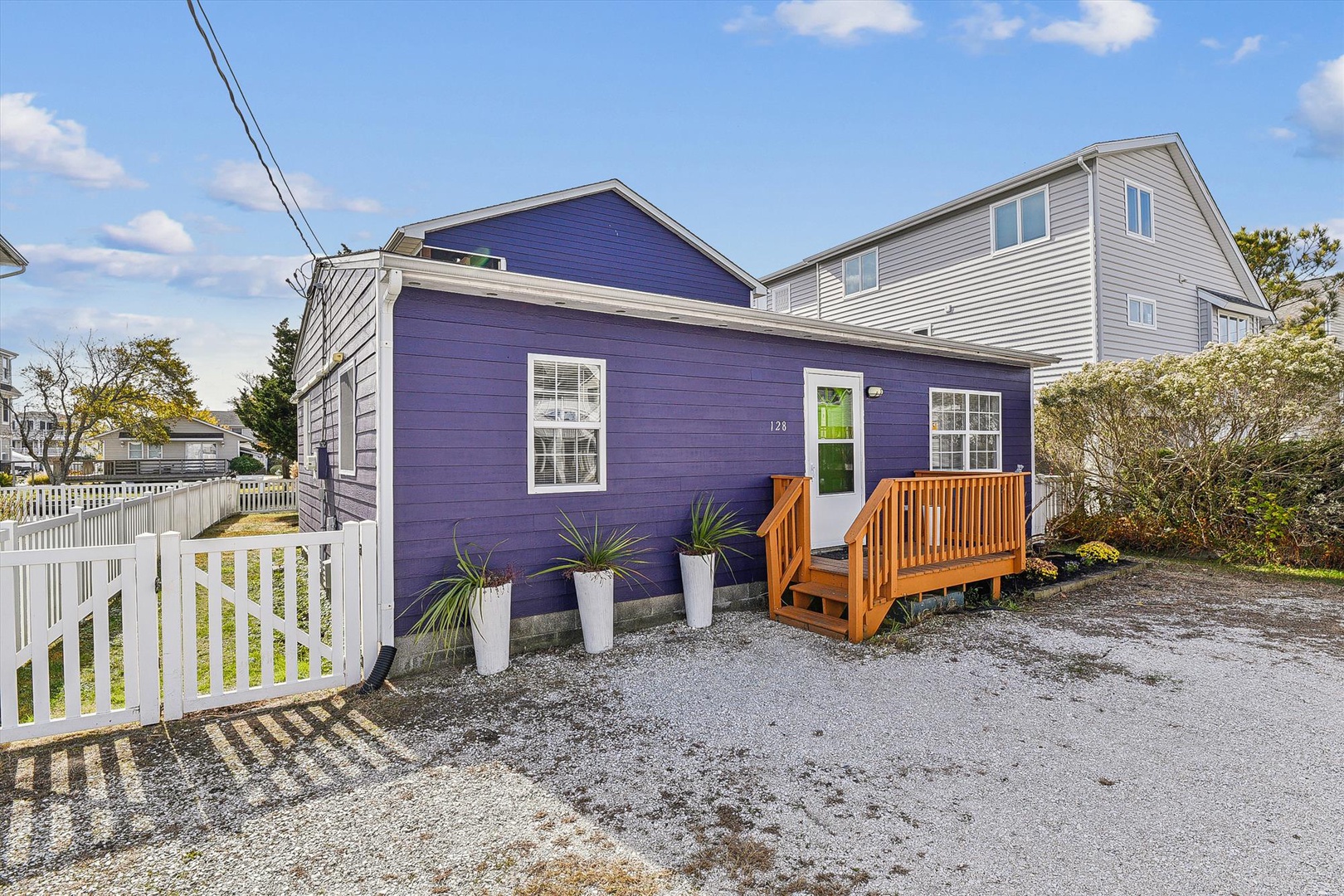 A vibrant purple cottage nestled in a peaceful residential neighborhood, featuring a welcoming entrance with modern landscaping and a sunny front patio perfect for coastal getaways.