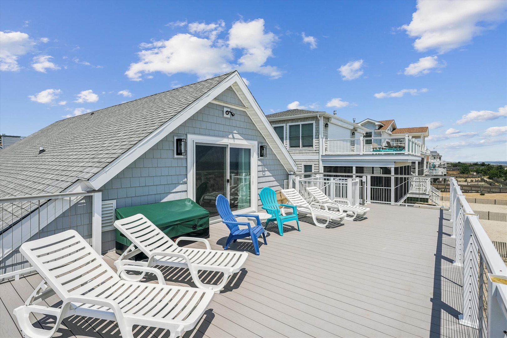 This rooftop deck sits atop a coastal property with beach access, offering an elevated outdoor lounging area with multiple sun chairs and Adirondack seating overlooking the neighborhood and beachfront area.