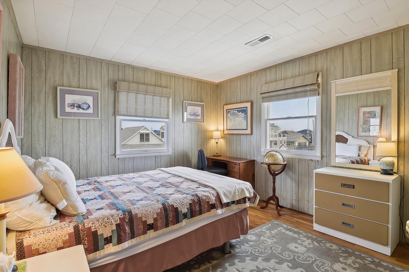 Drift off in this cozy bedroom featuring warm coastal charm, with soft natural light streaming through double windows, a handmade quilt, and thoughtful touches like a vintage globe and framed seaside art welcoming your restful escape.