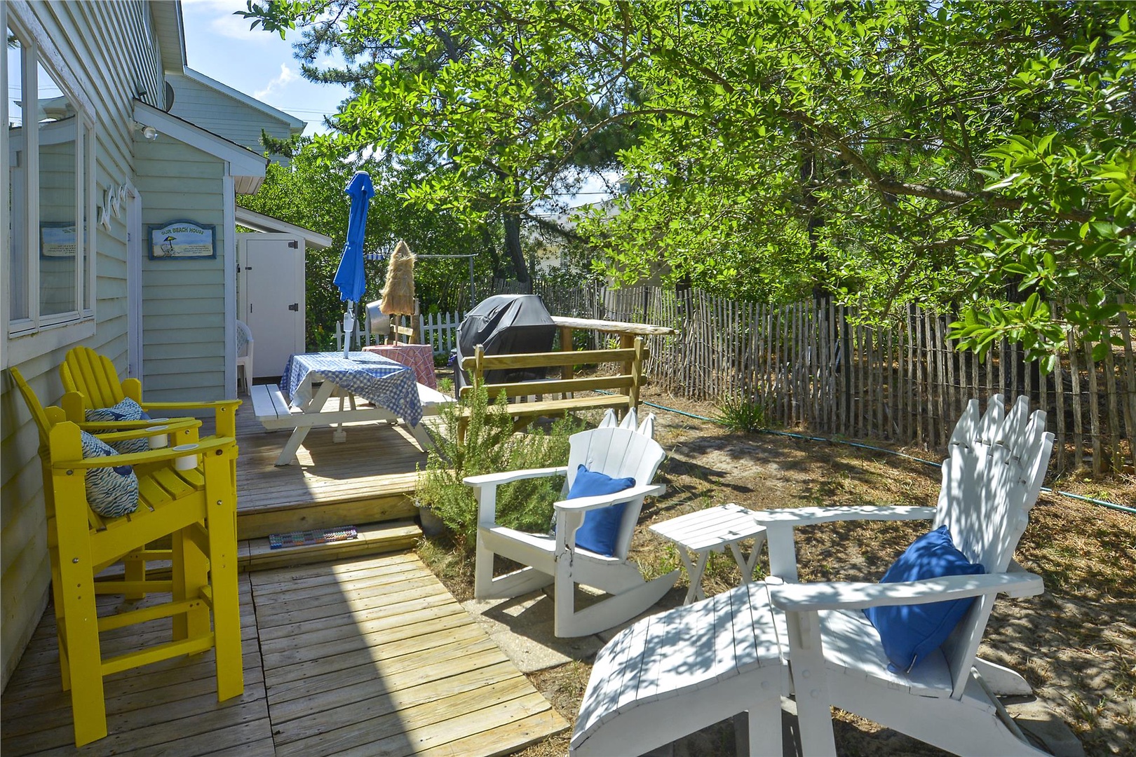 Private backyard retreat featuring sunny patio seating, outdoor dining area, and grassy yard bordered by mature trees and wooden privacy fencing—ideal for gathering or unwinding outdoors.