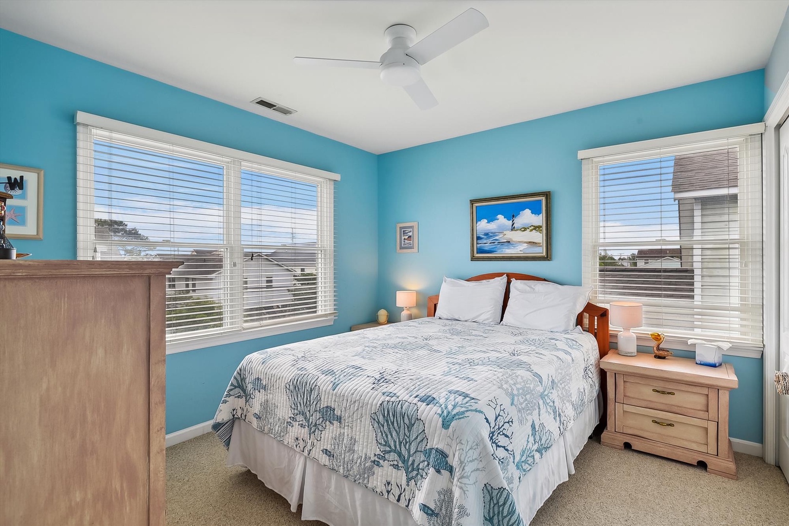 Your serene coastal bedroom features a plush queen bed, soothing ocean-blue walls, and ceiling fan to keep you comfortable. Bright windows invite natural light to fill your peaceful retreat.
