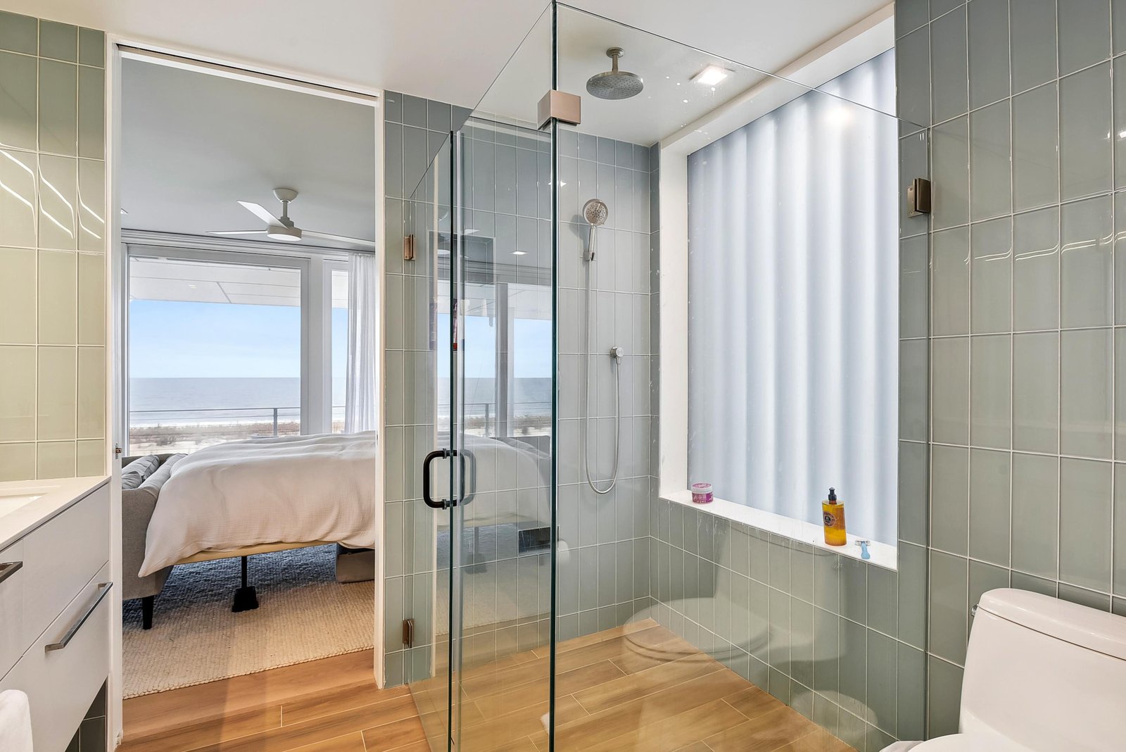 Step into a serene spa-like bathroom where soft green tiles and warm wood floors create your personal retreat. Your glass-enclosed shower overlooks the bedroom and beyond, with beach views just outside your window.