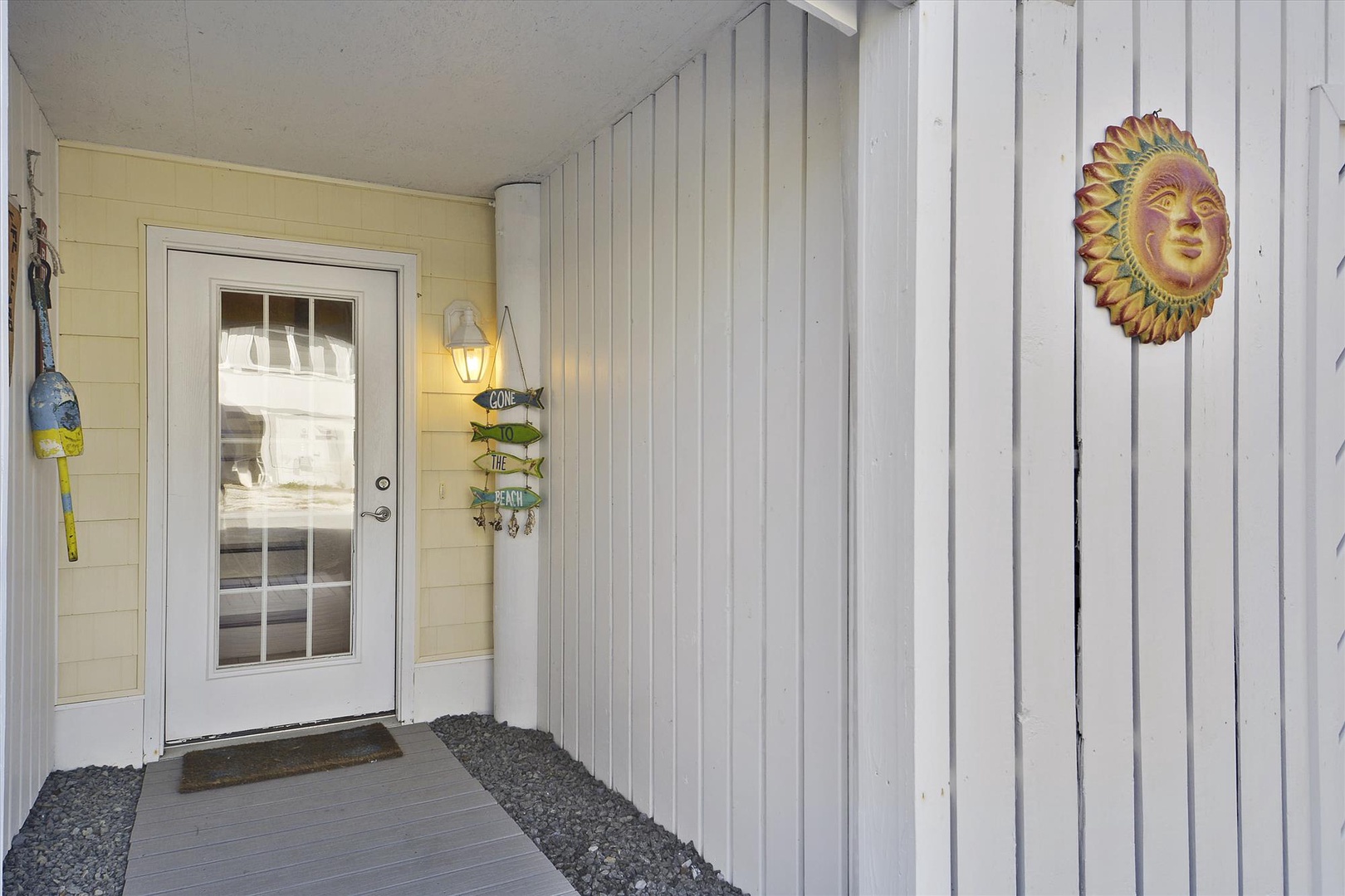 The entrance to the property features light-colored siding, a white door with glass panels, and decorative touches like a sun-face wall ornament and colorful fish accent, creating a welcoming arrival point.