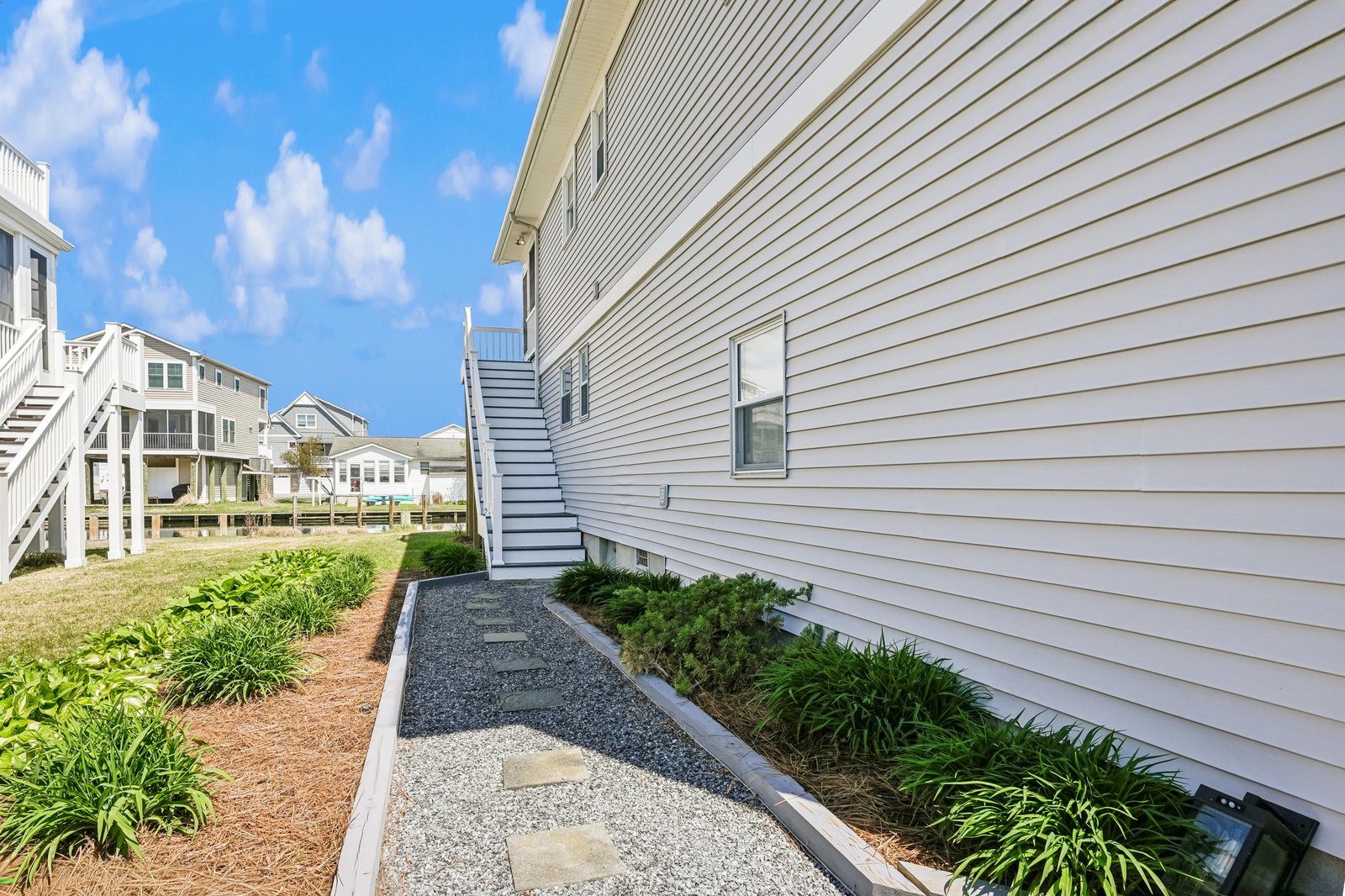 Coastal property walkway connecting neighboring beach houses with a private outdoor entrance featuring stepping stones and landscaped greenery under sunny blue skies.