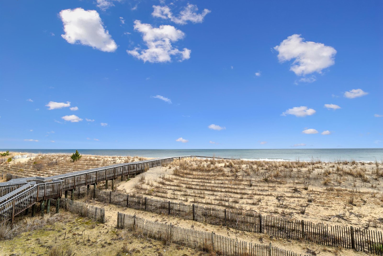 Oceanfront location with a peaceful sandy beach and protective dunes, framed by blue skies and calm waters that stretch to the horizon.