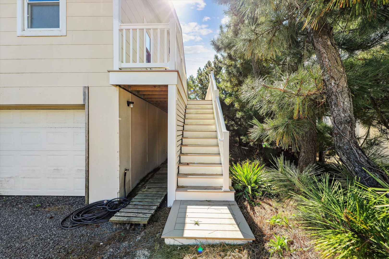 Coastal-style property entrance nestled among mature pines and tropical palms, featuring a private upper-level balcony and inviting outdoor stairway leading to your vacation retreat.