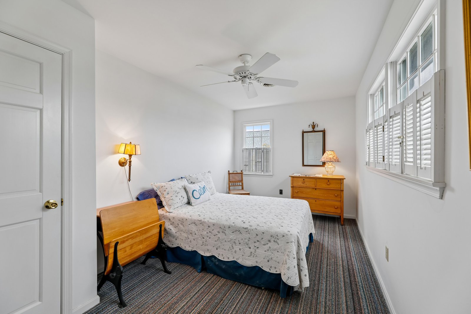 Your peaceful bedroom retreat awaits with crisp white walls, a cozy bed dressed in charming linens, and warm natural light streaming through shuttered windows. The ceiling fan keeps you cool while you unwind.