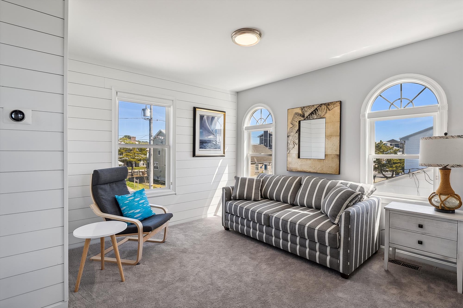 Settle into your bright coastal living space with shiplap walls and arched windows framing neighborhood views. Sink into the striped sofa, or curl up in the lounge chair—your serene retreat awaits.