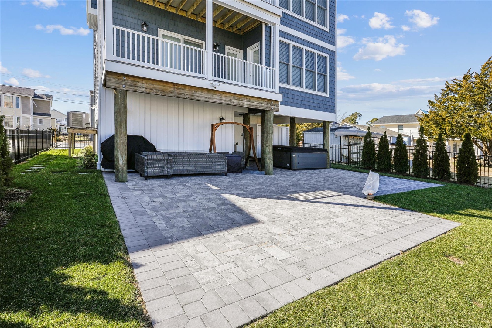 An elevated coastal-style property featuring a spacious paver patio beneath covered living areas, surrounded by manicured lawns and neighboring homes, photographed on a sunny afternoon.