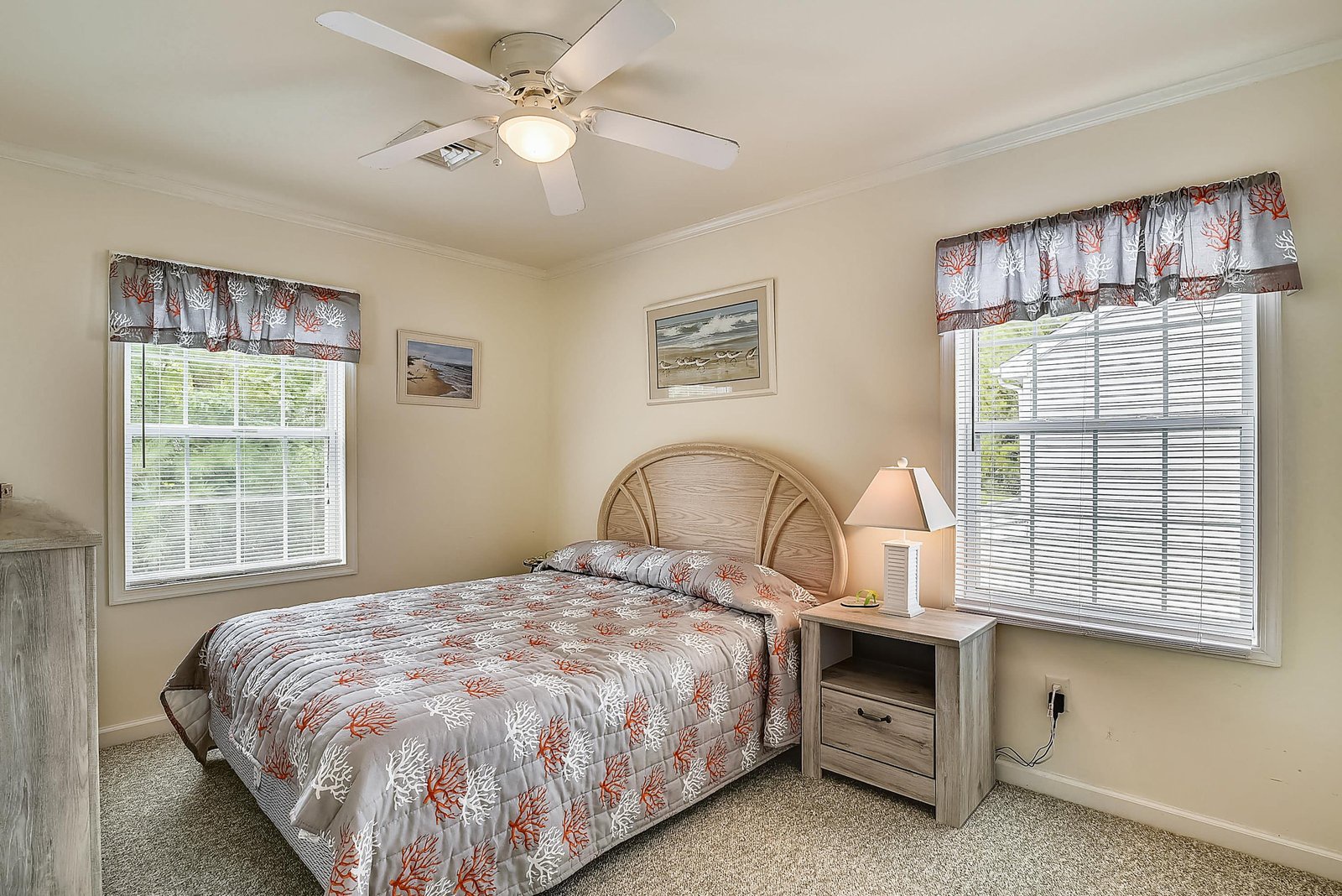 Your bedroom is a cozy retreat with soft coastal touches, plush carpeting, and a ceiling fan for your comfort. Natural light fills the space through large windows with charming valances and peaceful garden views.