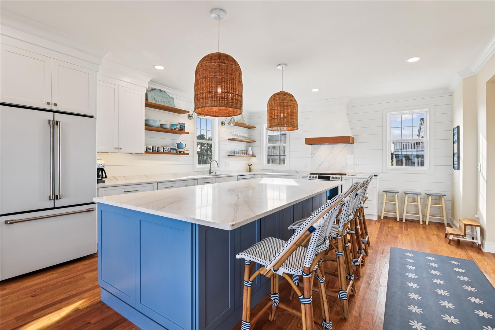 Bright coastal-inspired kitchen with marble island, warm woven pendant lights, and charming bar seating. Your mornings begin here with coffee and natural light streaming through shiplap walls.