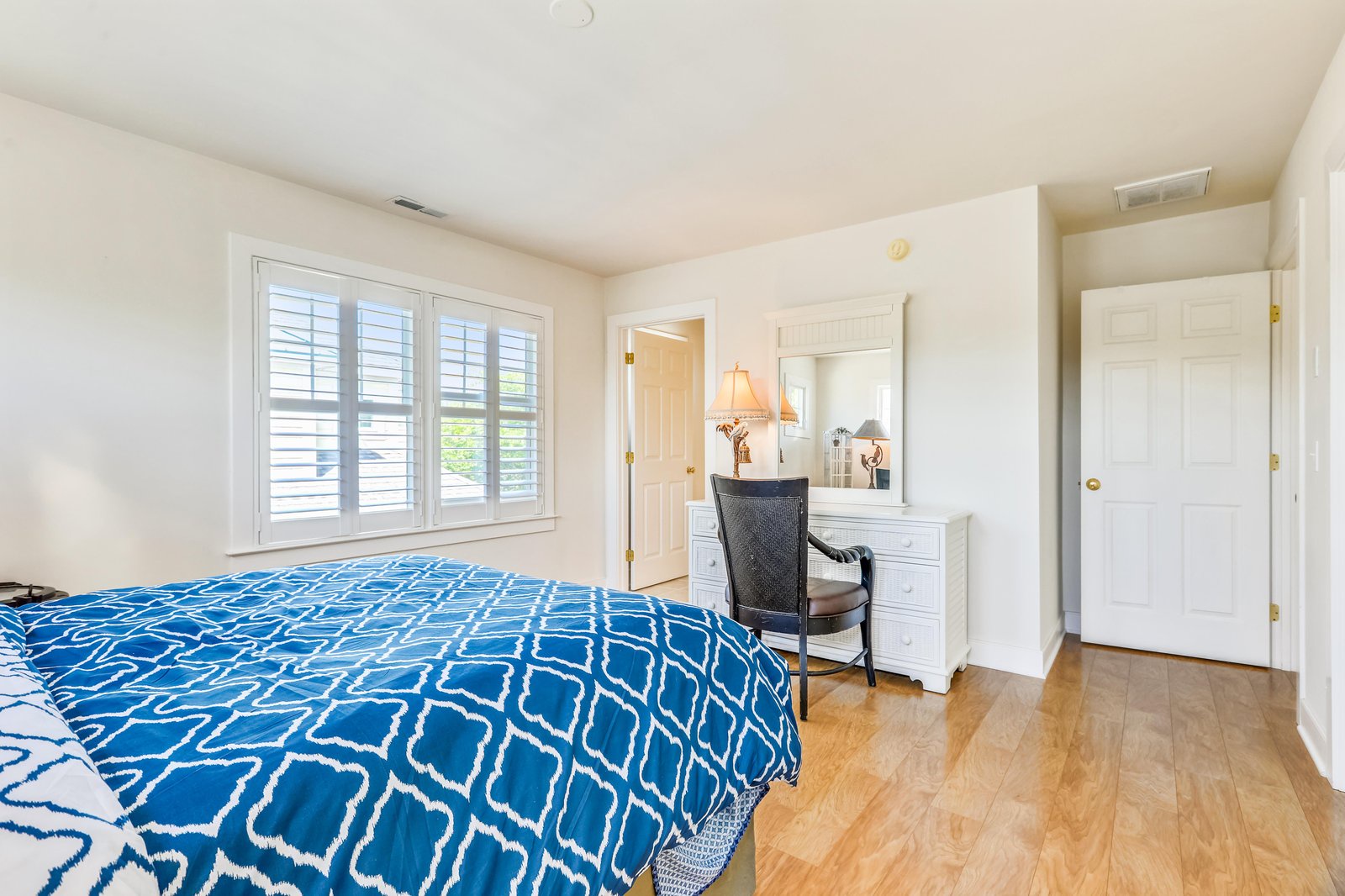 Relax in your bright, peaceful bedroom with warm wood floors, natural light from plantation shutters, and a vanity ready for your morning routine.