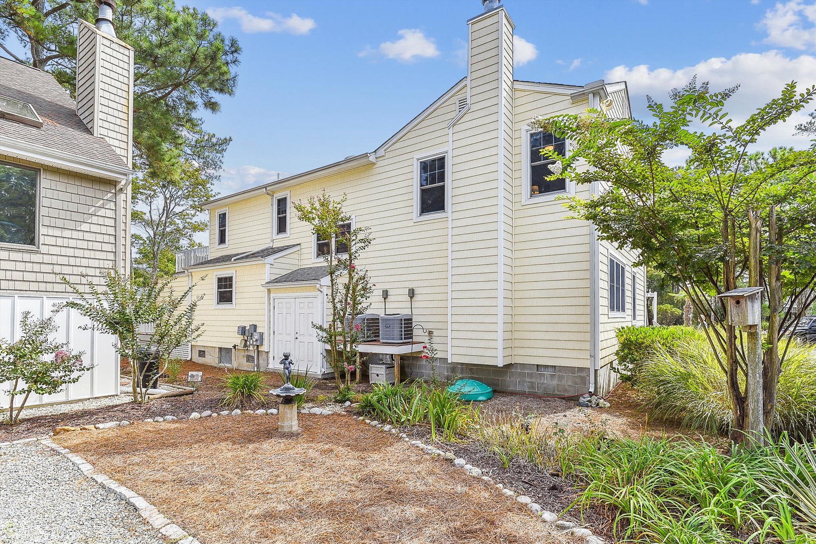 A charming two-story vacation rental surrounded by mature trees and established landscaping. The property features multiple windows, a well-maintained exterior, and easy parking access.