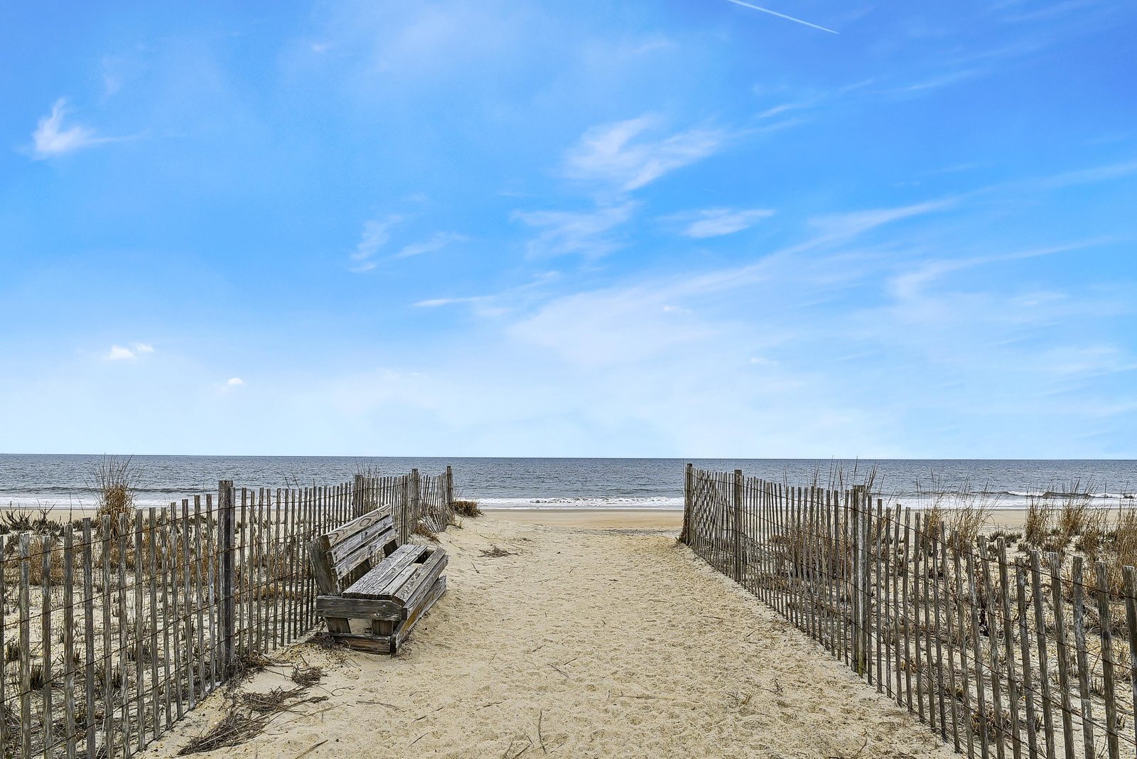 A peaceful beach boardwalk with a weathered bench invites rest before sandy shores stretch to meet calm, sparkling waters under soft blue skies.