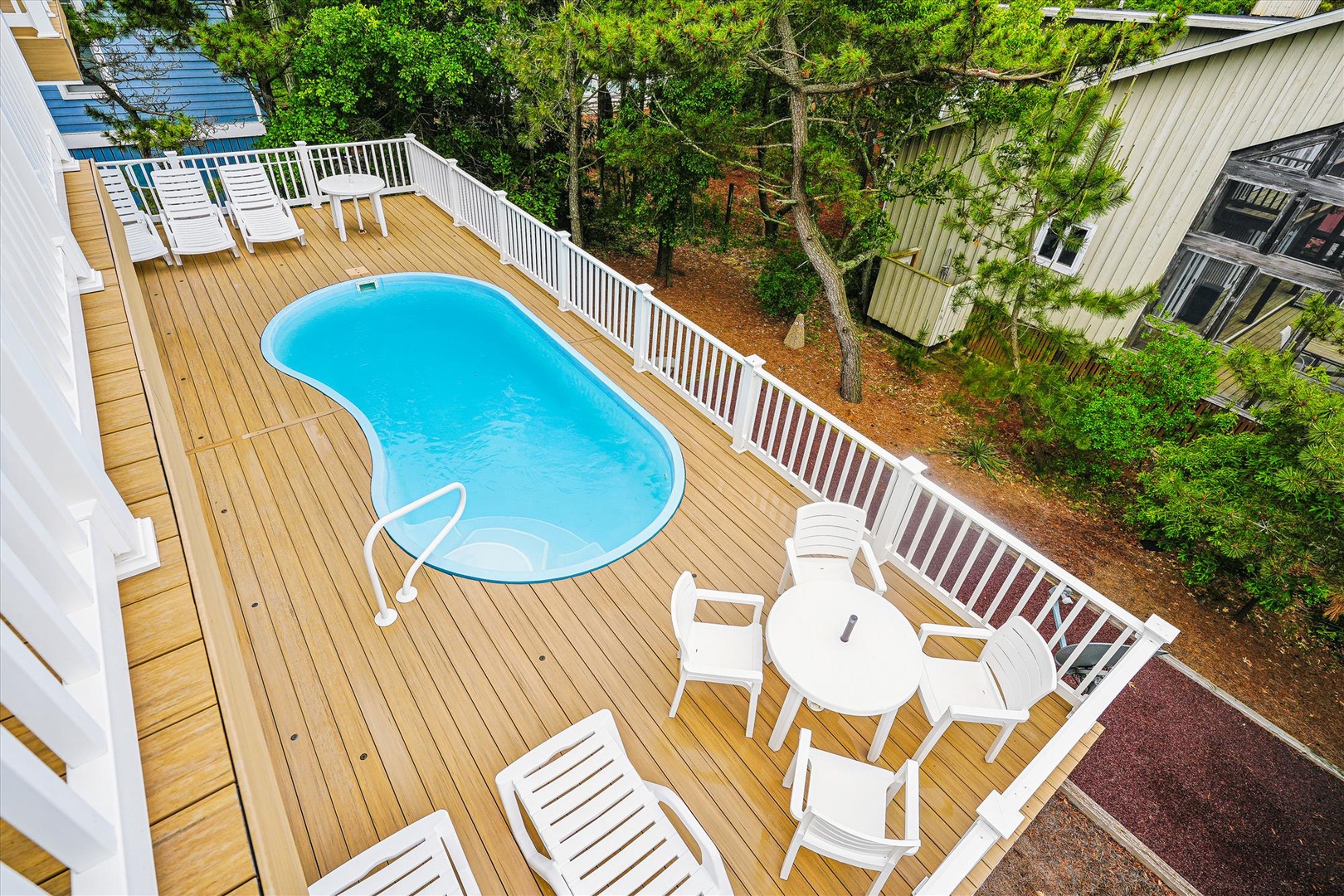 An elevated view shows the spacious wooden deck surrounding the private pool, furnished with white lounge chairs and a dining set, nestled among lush trees.