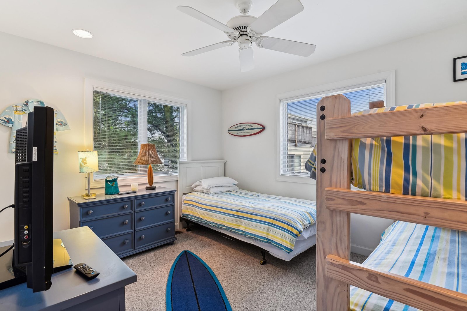 Beach-themed bedroom with bunk and twin beds, cheerful striped linens, ceiling fan, and natural light. Surf-inspired décor creates a relaxed coastal vibe perfect for unwinding.